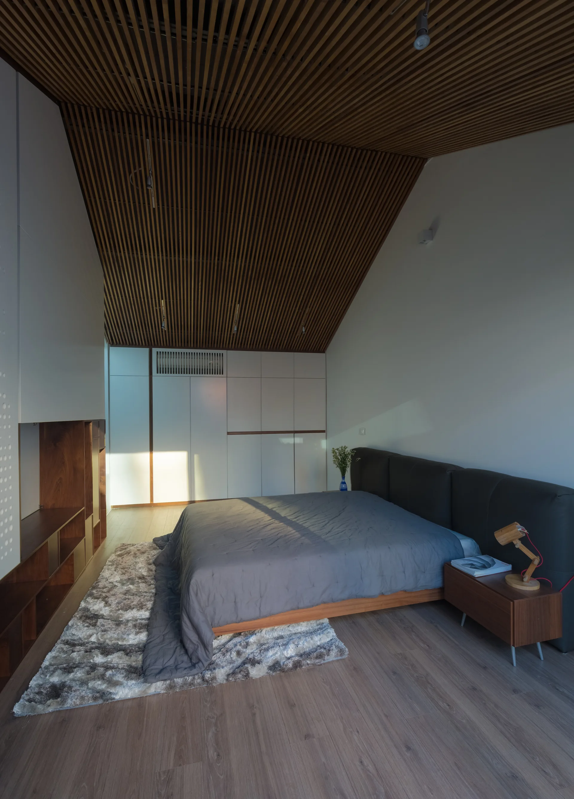 A contemporary bedroom features a low-profile bed, built-in white wardrobes, and a sloped ceiling lined with parallel wooden slats.