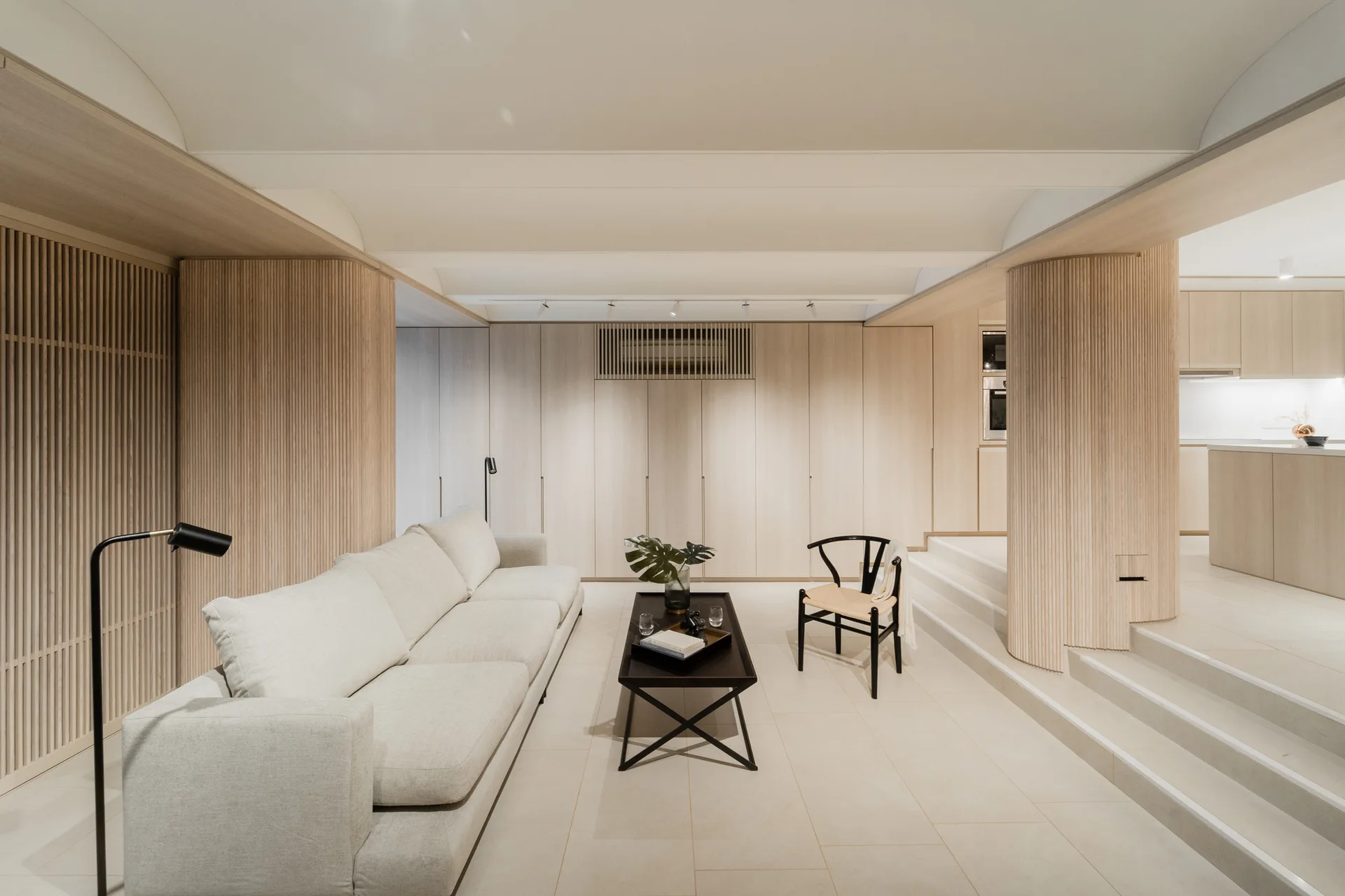 A sunken minimalist living room featuring an off-white sofa, vertical wood slat walls, and steps leading to an elevated kitchen area.