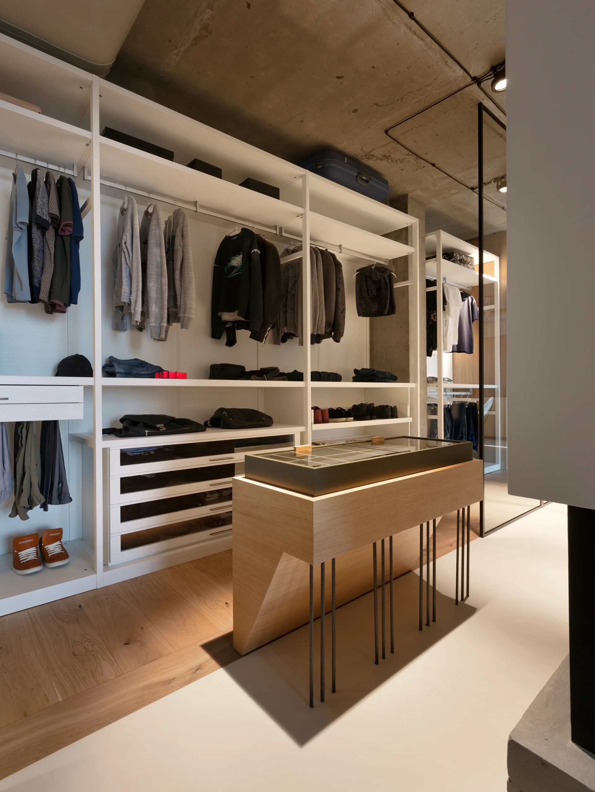 A walk-in closet featuring white metal open shelving, a central light wood display island with a glass top, and an exposed raw concrete ceiling.