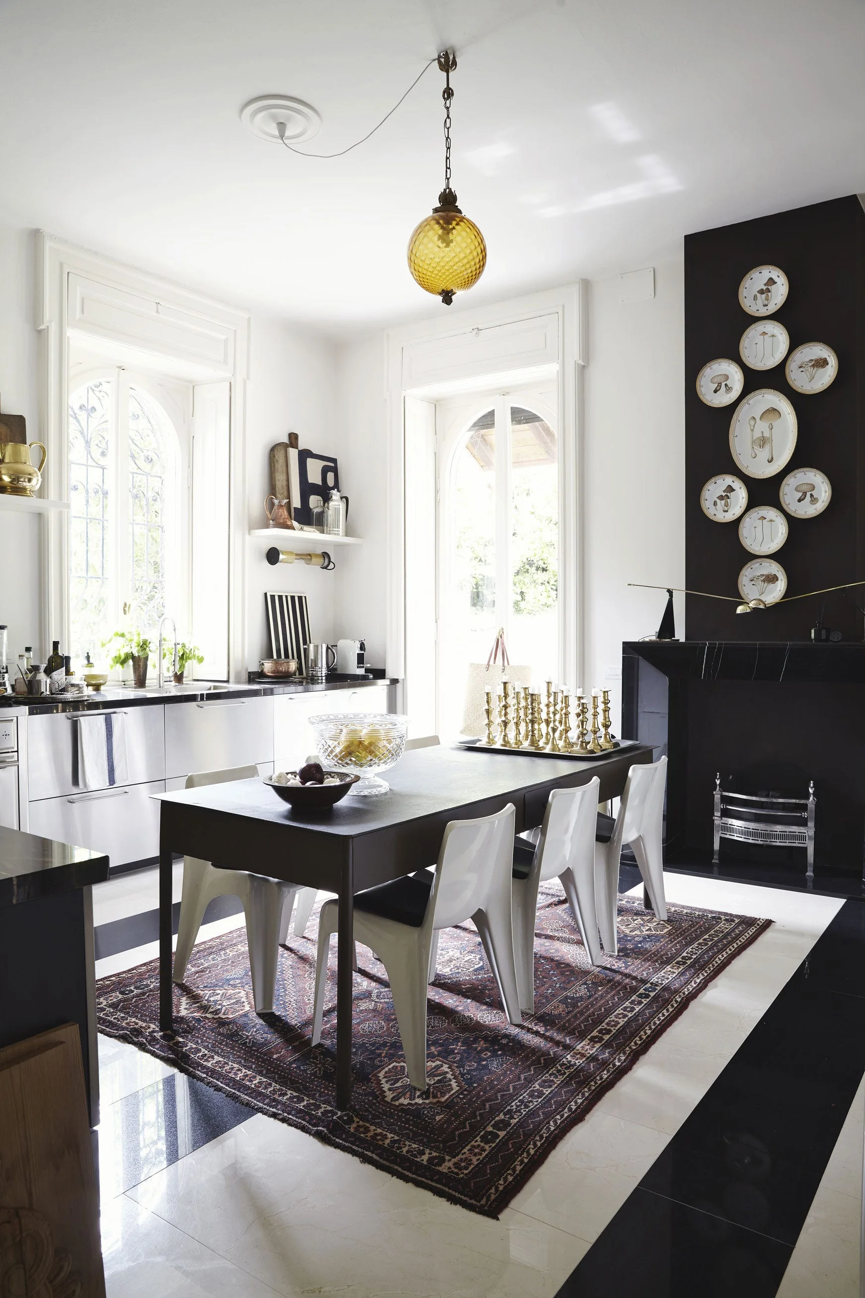 A modern dining room features a dark table on an ornate rug, next to a black fireplace with a vertical plate collection.
