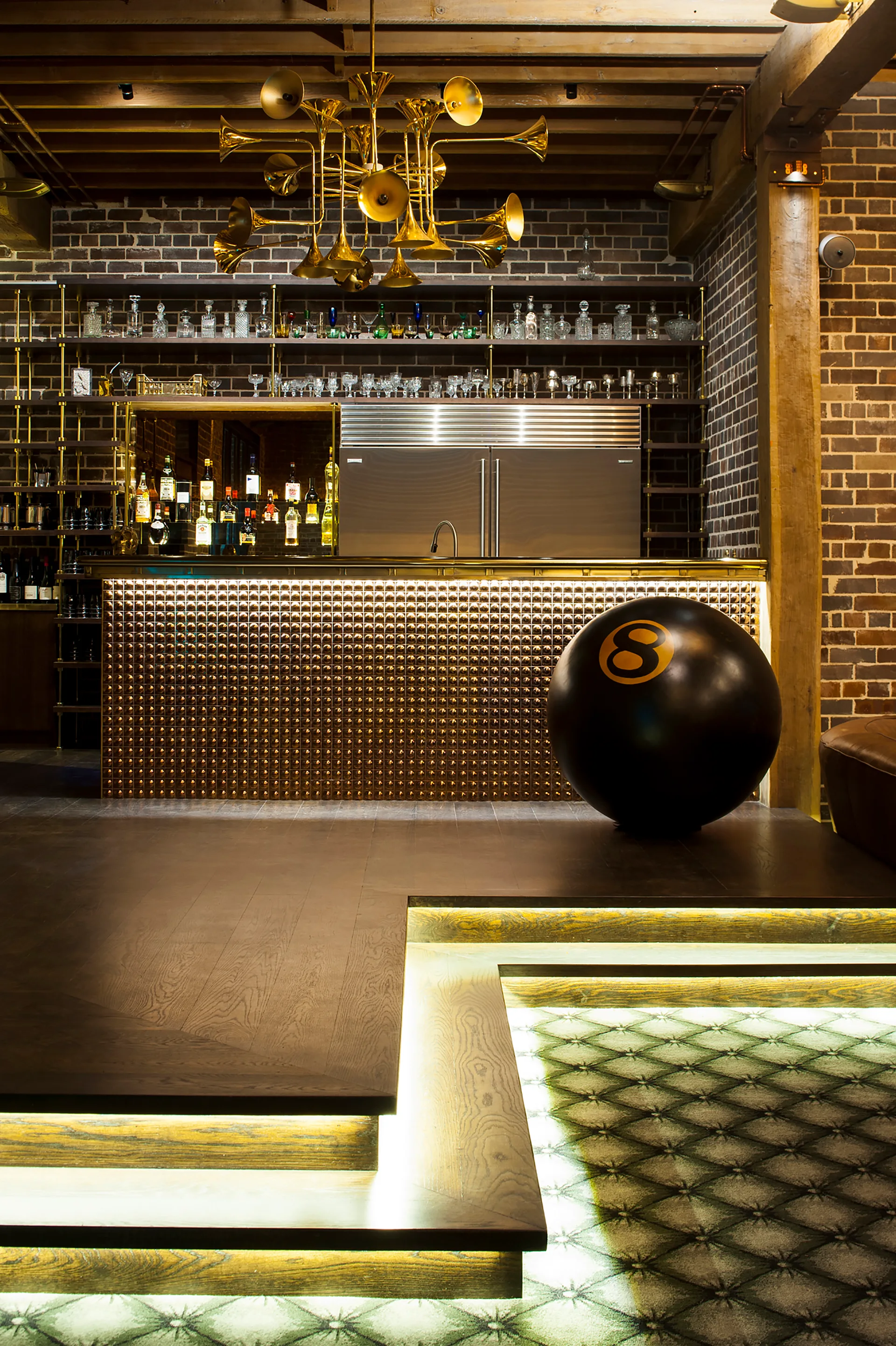 An industrial bar area features a textured brass counter, a trumpet chandelier, exposed brick walls, and a tiered platform with integrated step lighting.