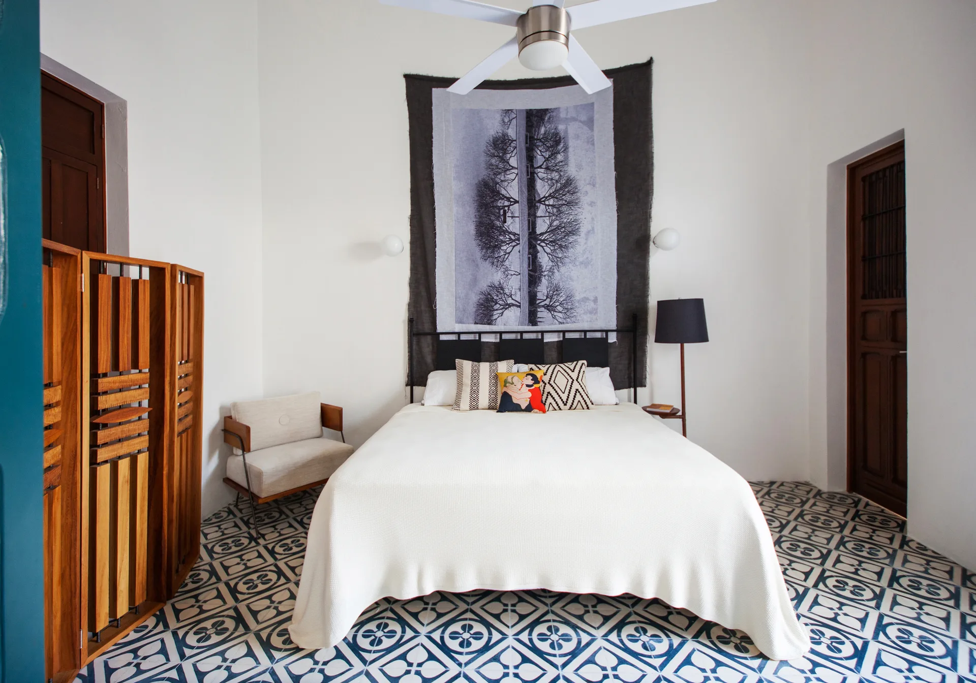 An eclectic bedroom features a white covered bed backed by a large photographic tapestry, with geometric patterned floor tiles and a wood screen.