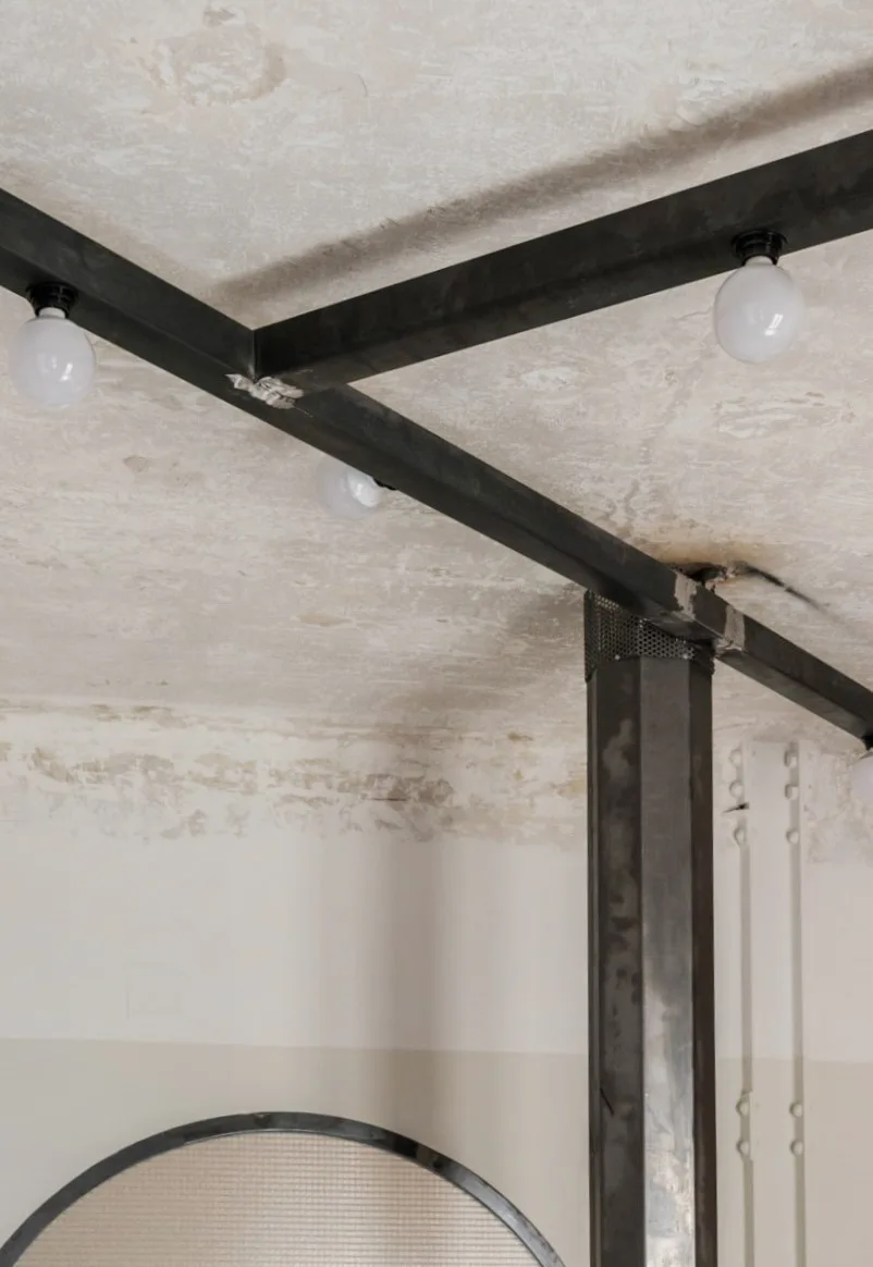 An exposed concrete ceiling features intersecting black steel beams with mounted white globe bulbs and a matching metal support column.