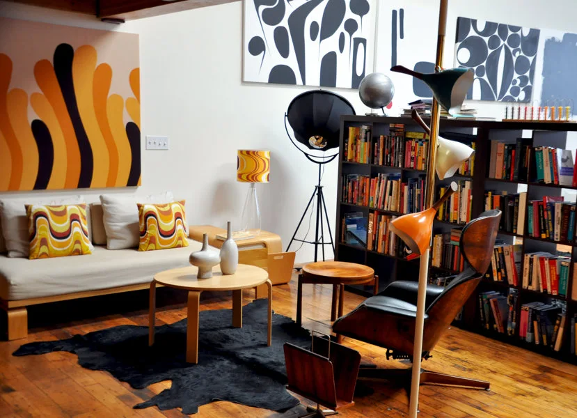 A retro living room featuring a low sofa with patterned orange pillows, a black cowhide rug, a dark wood bookcase, and abstract wall art.
