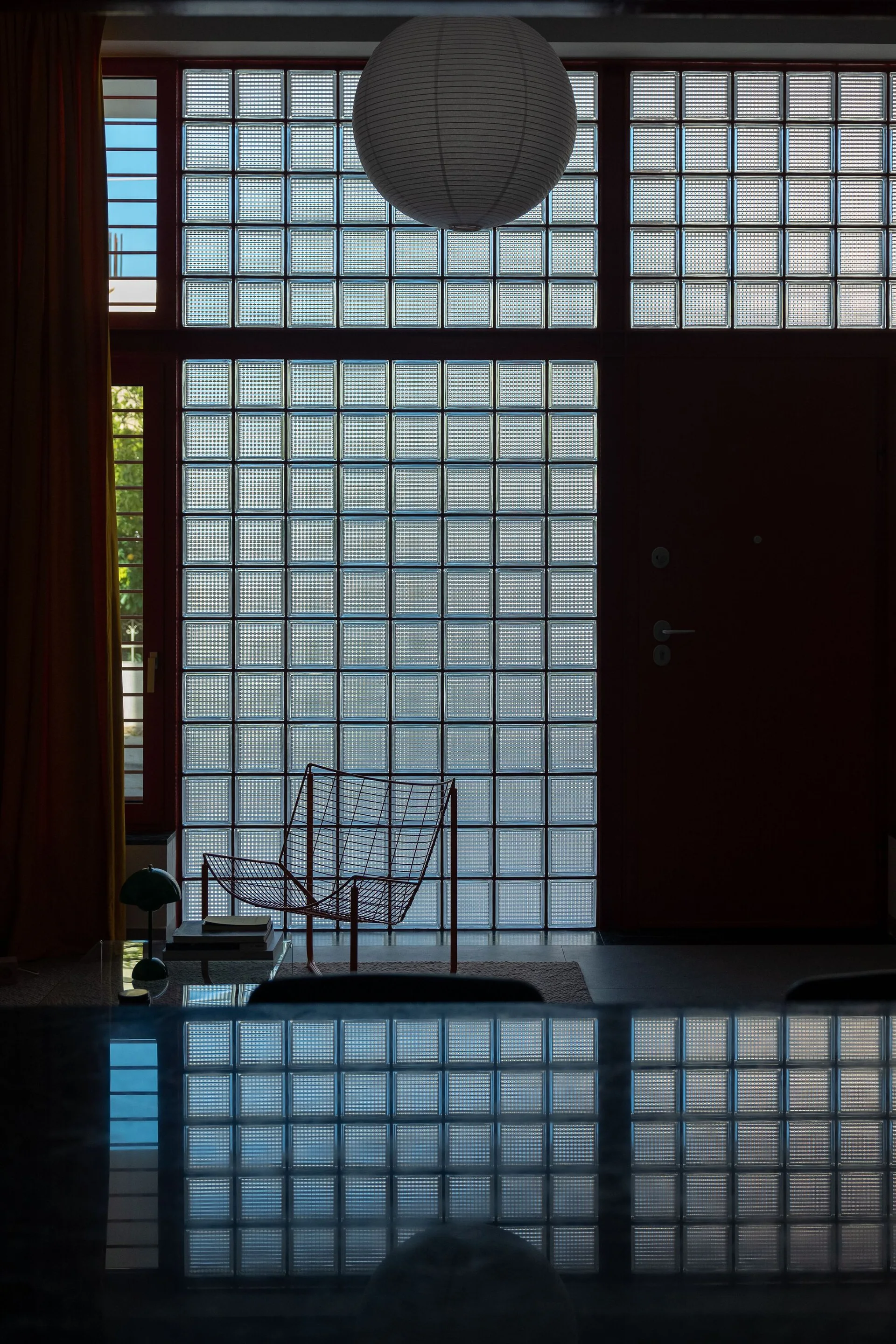 A red wire accent chair sits in front of a large grid wall made of translucent glass blocks under a paper globe pendant light.