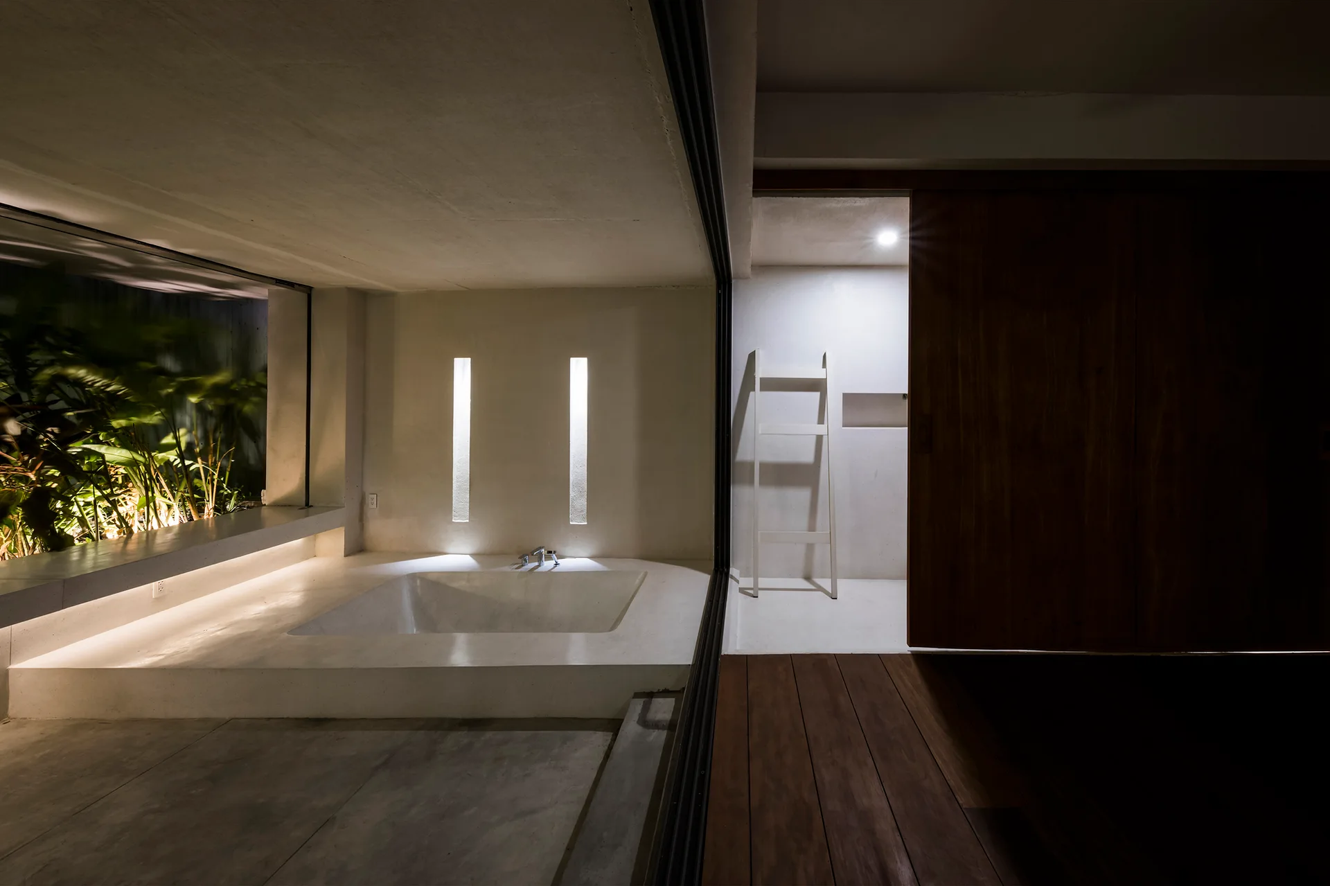 A minimalist bathroom with a built-in sunken concrete soaking tub, vertical wall lights, and a dark wooden sliding door.