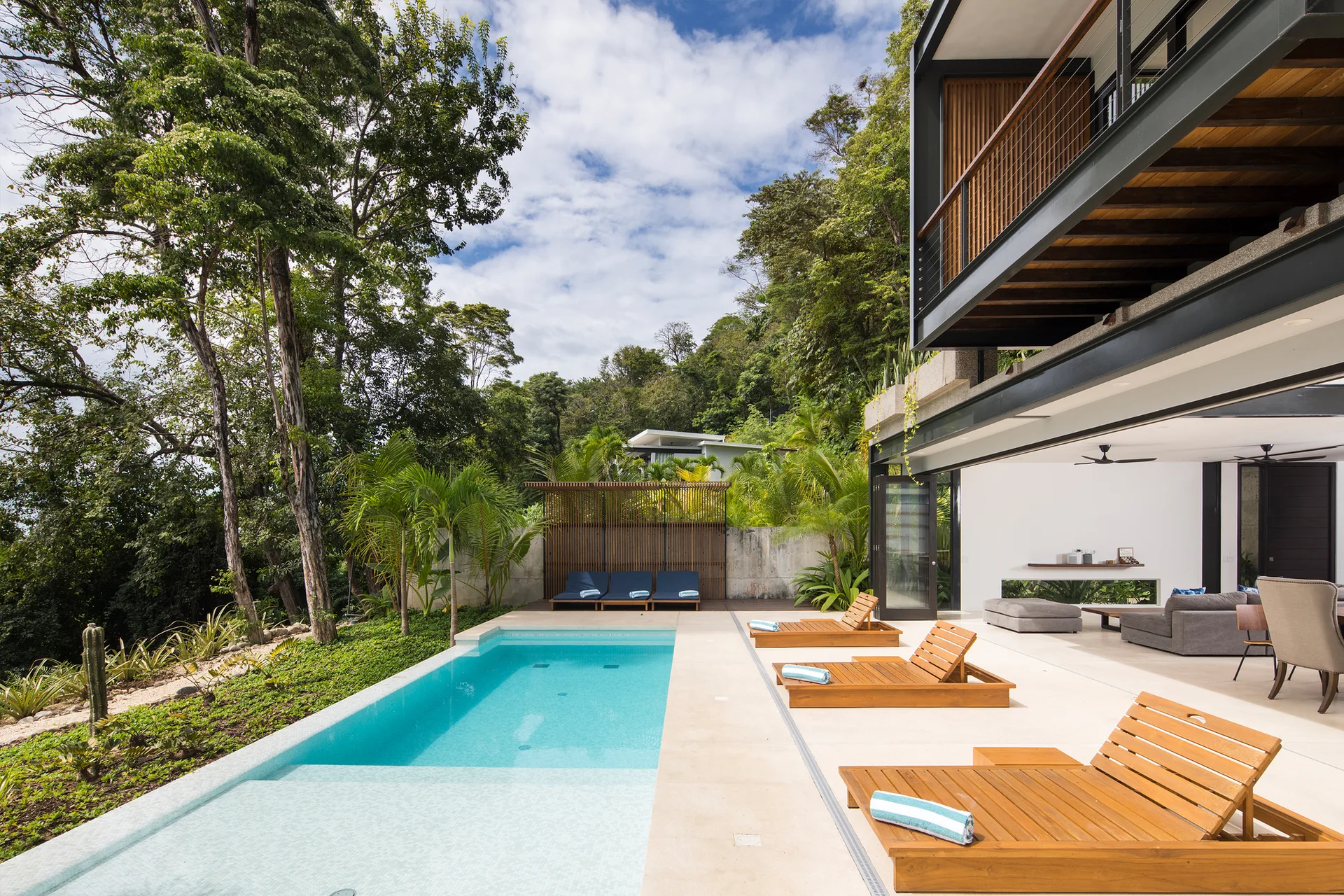 A rectangular swimming pool with wooden lounge chairs sits next to an open-air modern living room surrounded by lush tropical forest.