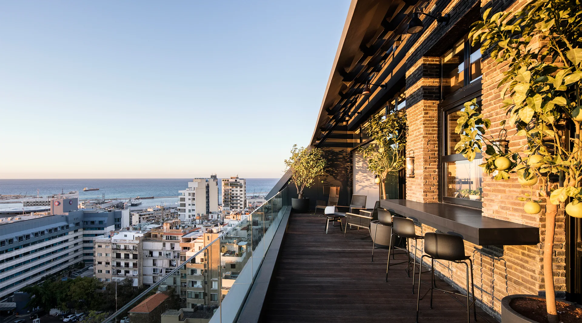 A rooftop terrace features a dark brick wall with a black bar counter, black bar stools, potted trees, and a glass railing overlooking the city skyline.