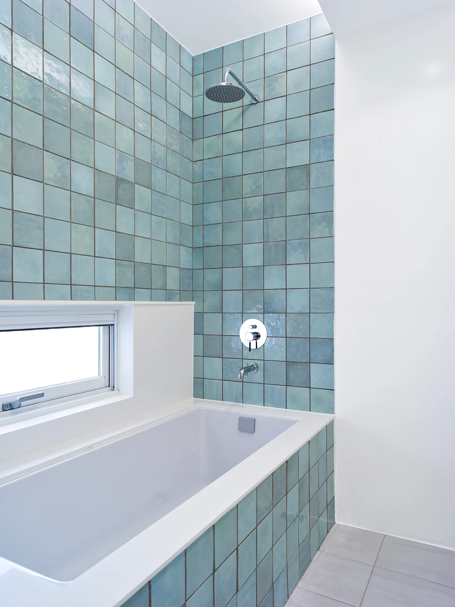 A modern bathroom features a white alcove bathtub surrounded by glossy teal blue square wall tiles and chrome shower fixtures.
