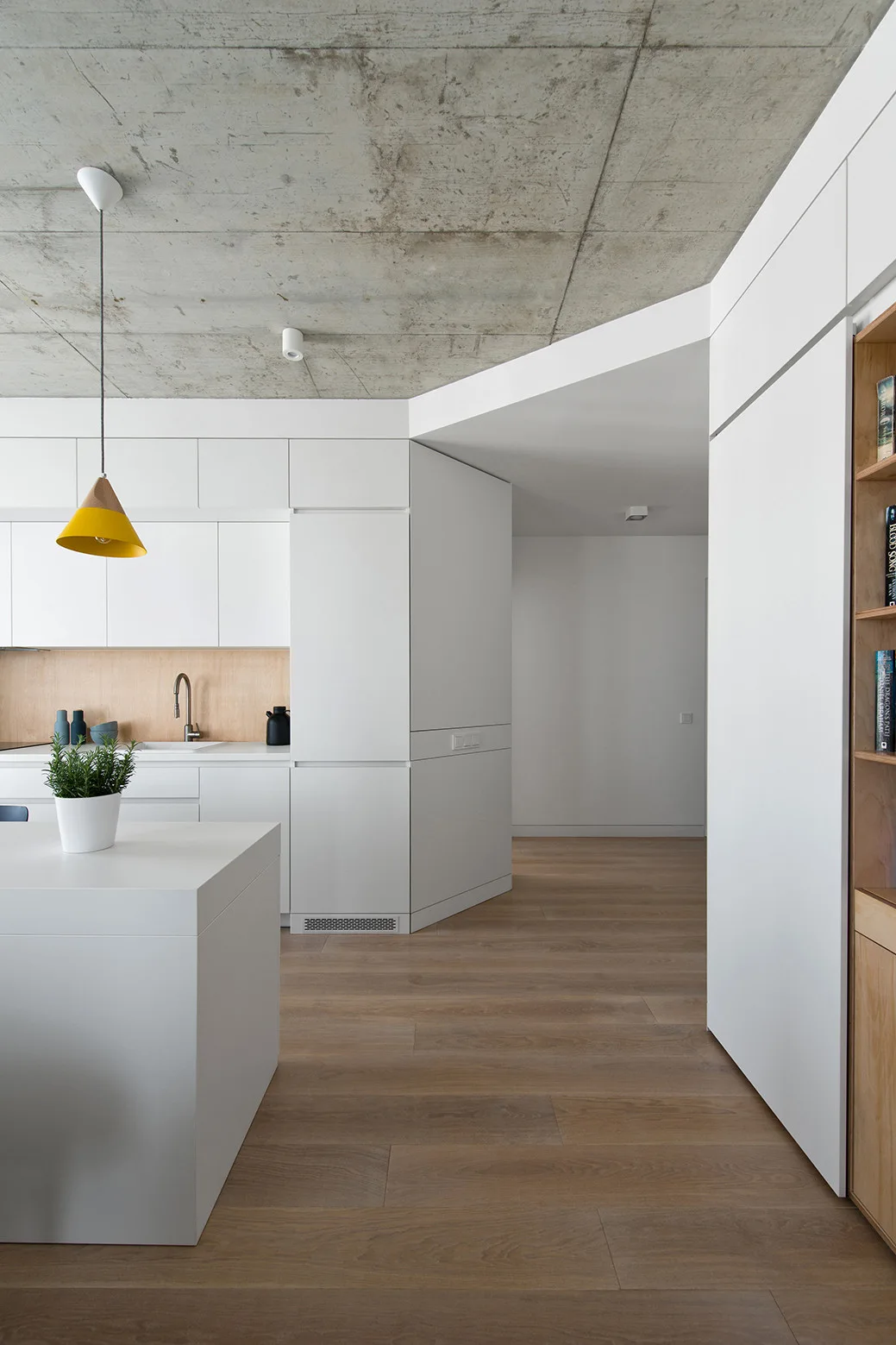 A minimalist kitchen features white handleless cabinetry, a light wood backsplash, and a yellow pendant light under an exposed concrete ceiling.