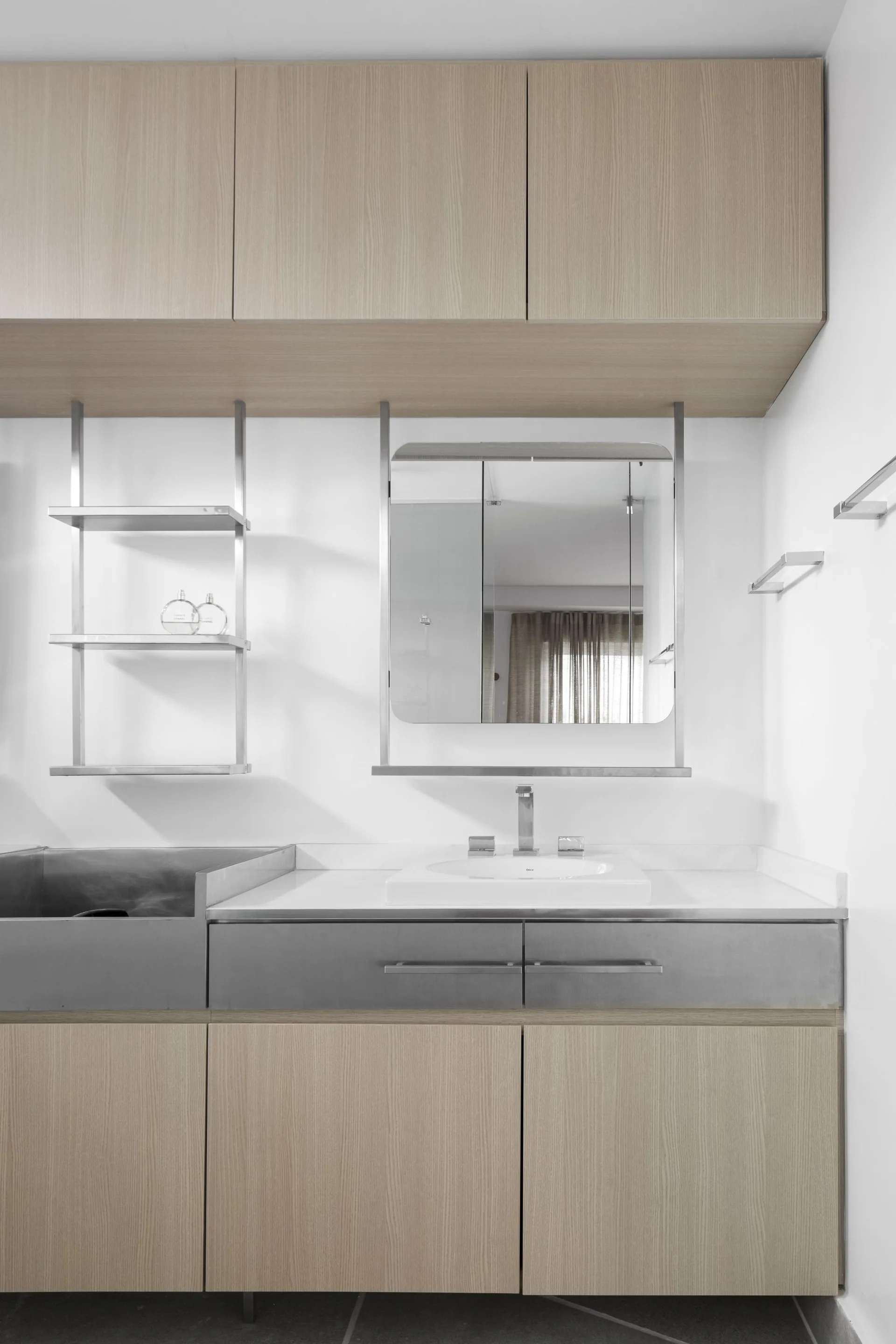 A modern bathroom vanity featuring light wood cabinets, a white undermount sink, and metal shelving suspended beneath the upper cabinetry.