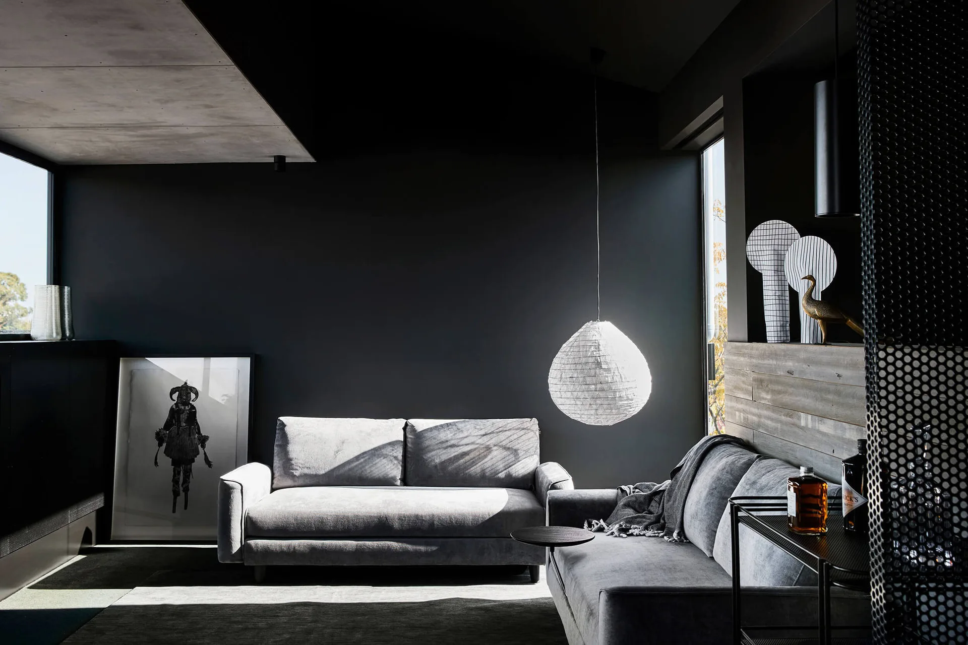 A moody dark grey living room features two grey upholstered sofas, a large white textured paper pendant light, and a framed black and white illustration.