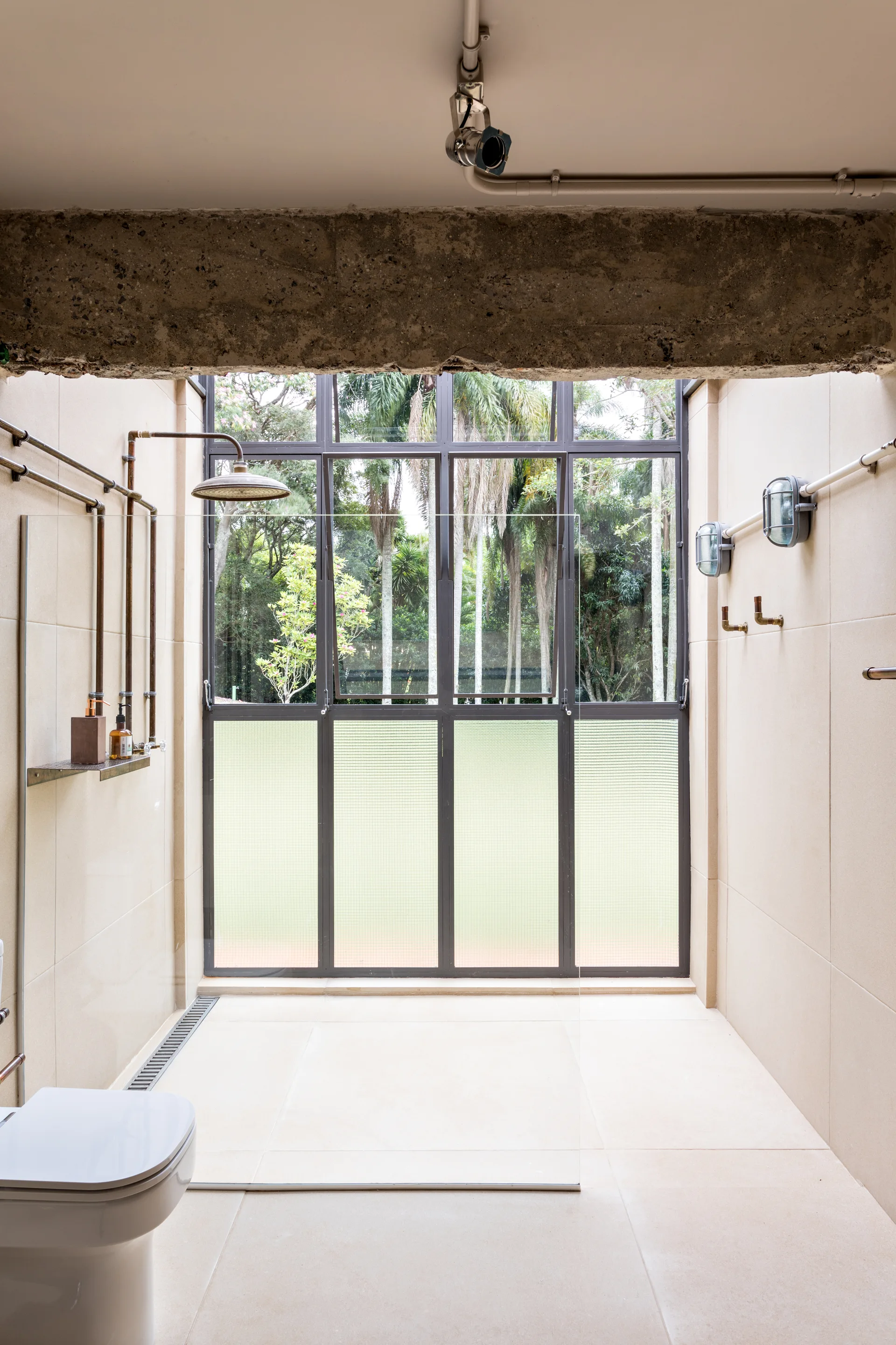 An industrial bathroom features a tiled shower with exposed copper plumbing, a glass partition, and a large window looking out onto a garden.