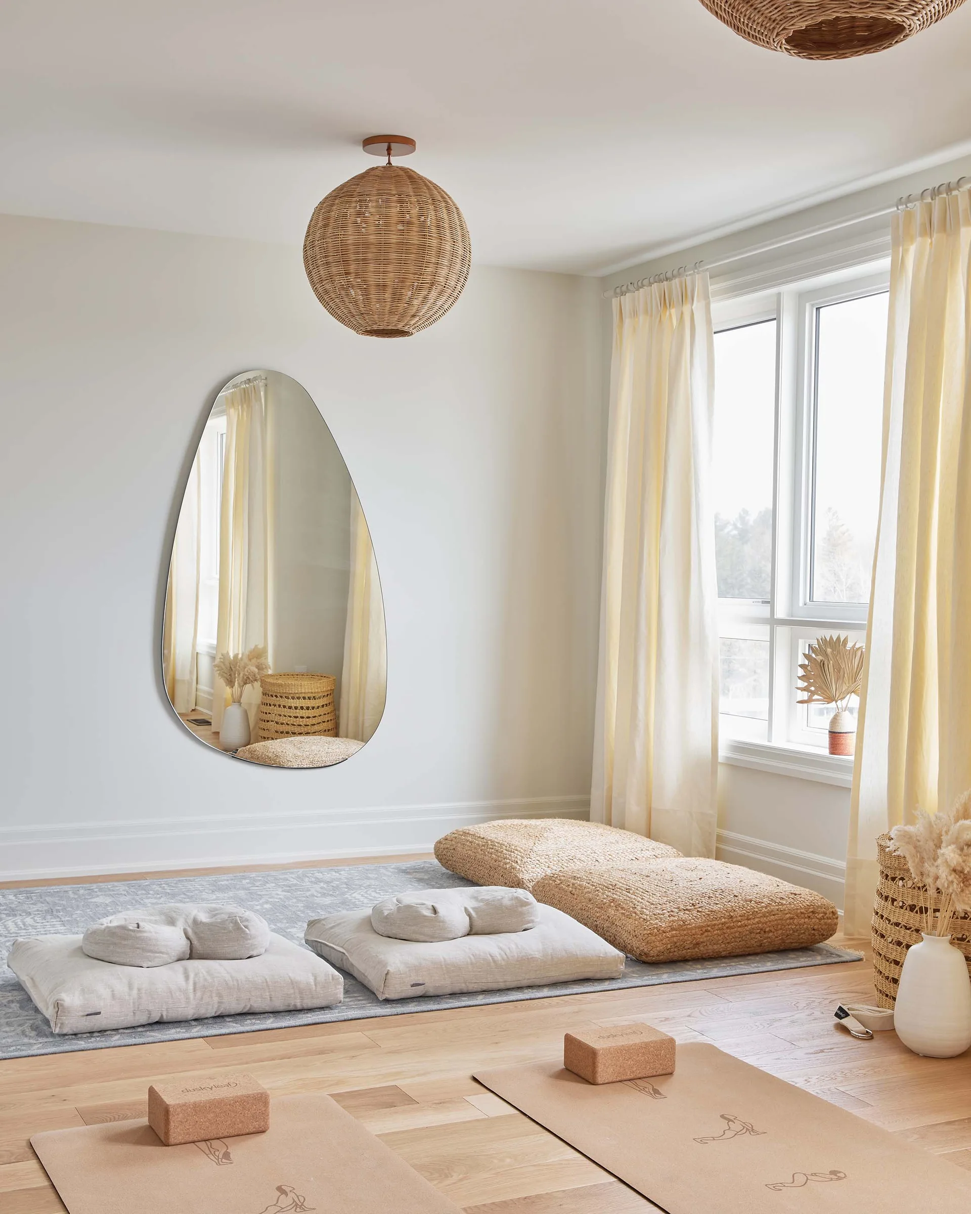 A bright meditation room features linen floor cushions, yoga mats on light wood floors, and a large asymmetrical mirror on an off-white wall.