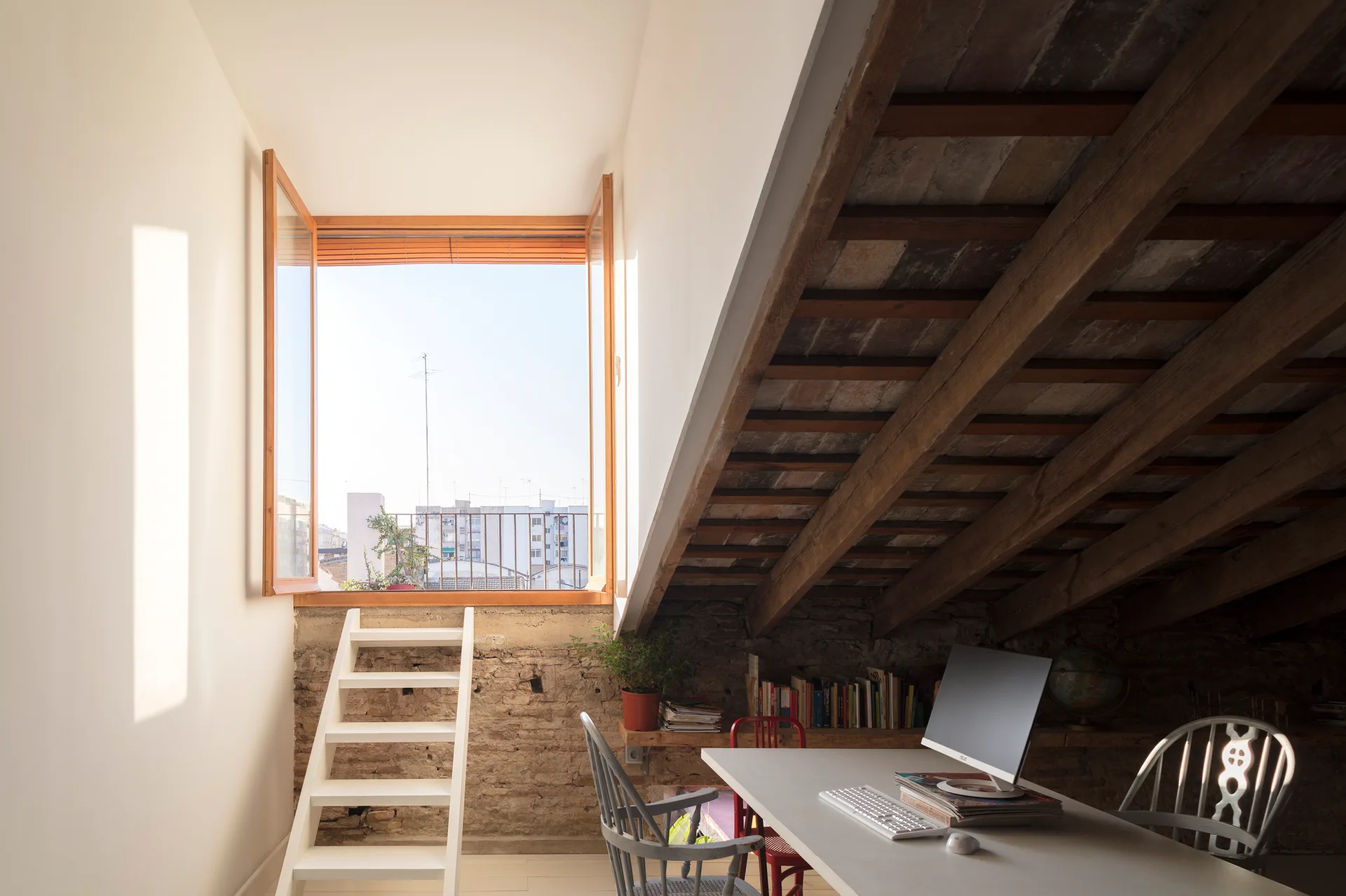 A rustic attic workspace featuring a white desk, exposed brick, sloped timber beams, and a white ladder leading to an open window.