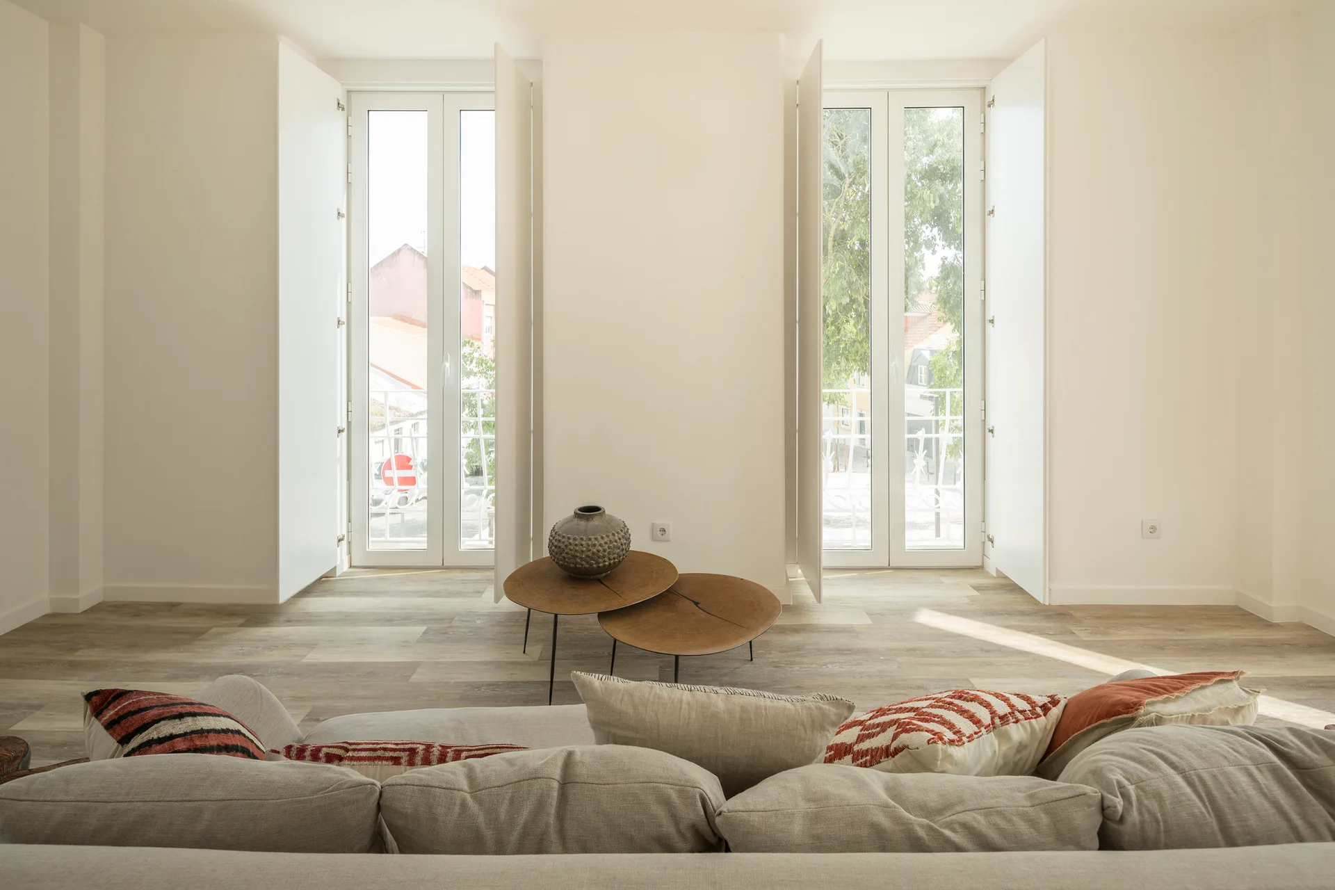 A minimalist living room featuring a grey fabric sofa, nested wooden coffee tables, and two tall white shuttered windows.