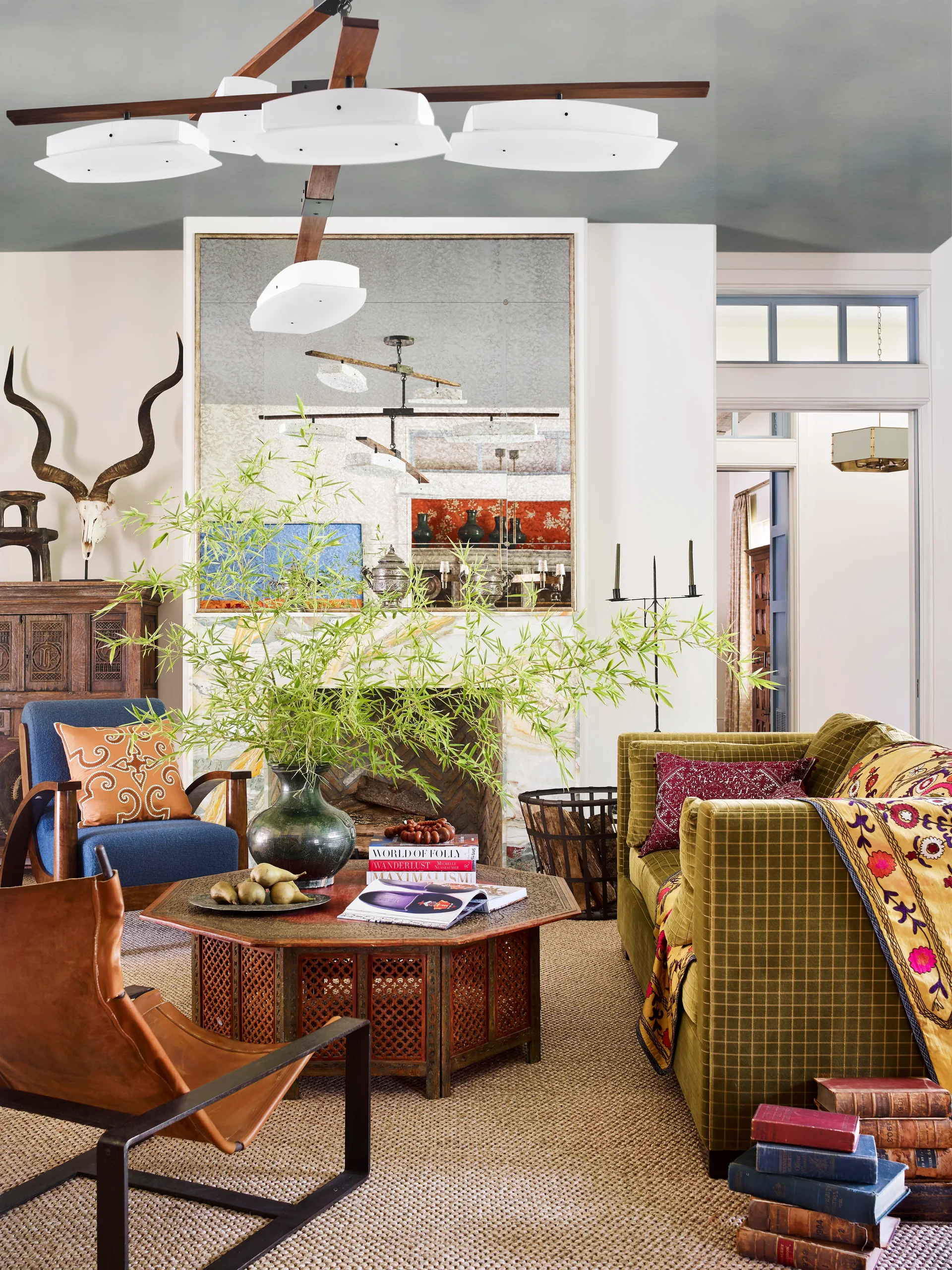 An eclectic living room features an olive green checkered sofa, an octagonal carved wooden coffee table, and an antique mirror above a fireplace.