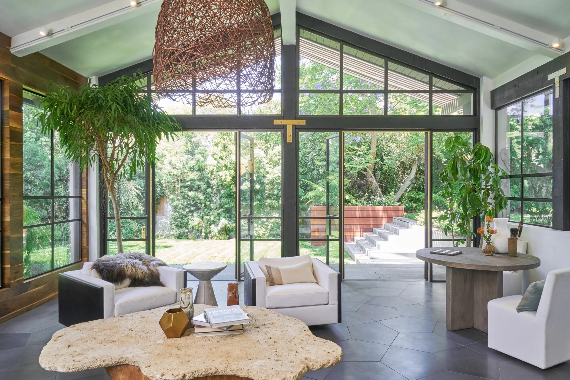 A sunlit living room features white armchairs, a raw stone coffee table, a large woven pendant light, and a floor-to-ceiling glass wall opening to a garden.