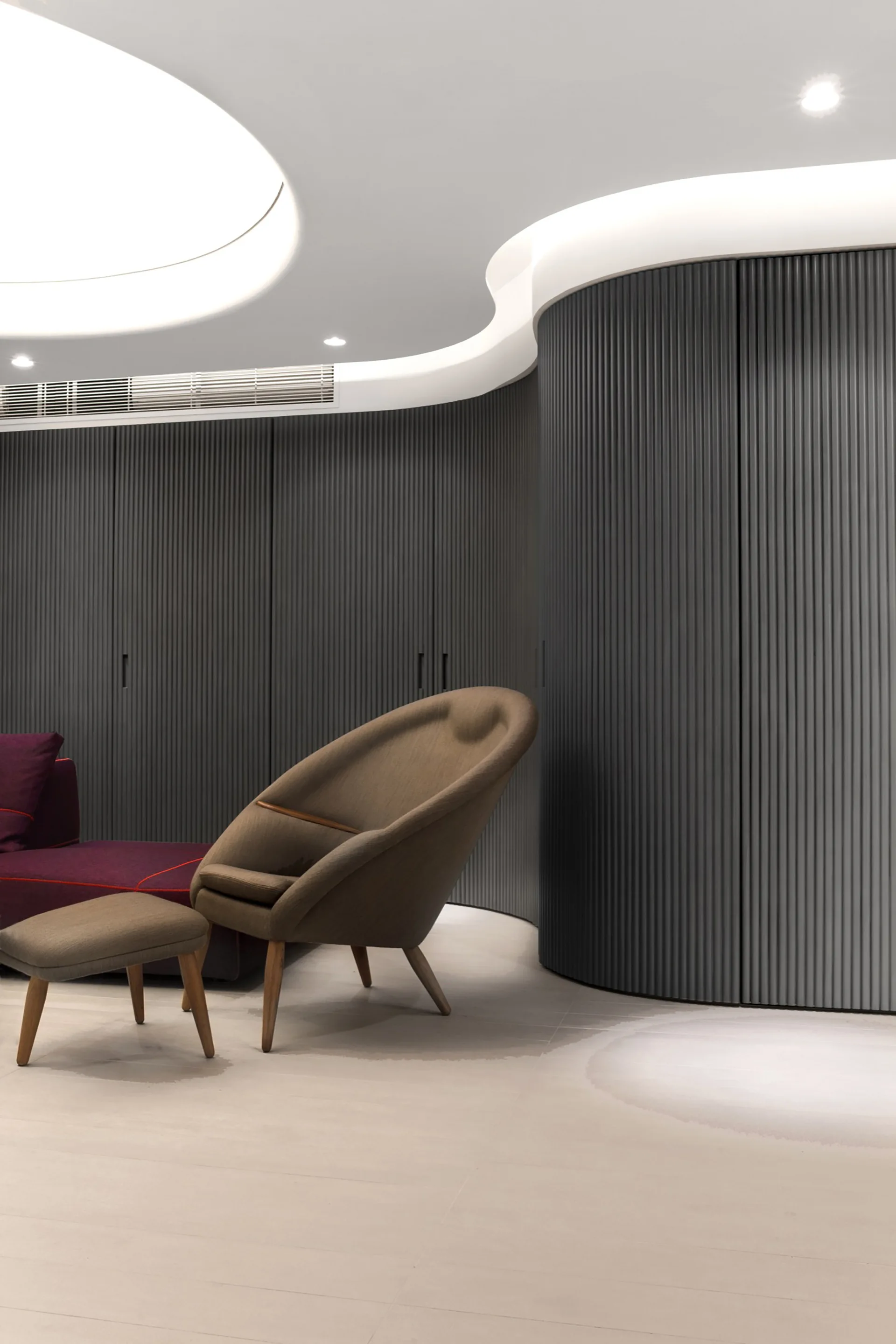 A contemporary living space with a brown upholstered lounge chair and ottoman set against a dark grey curved fluted wall with cove ceiling lighting.