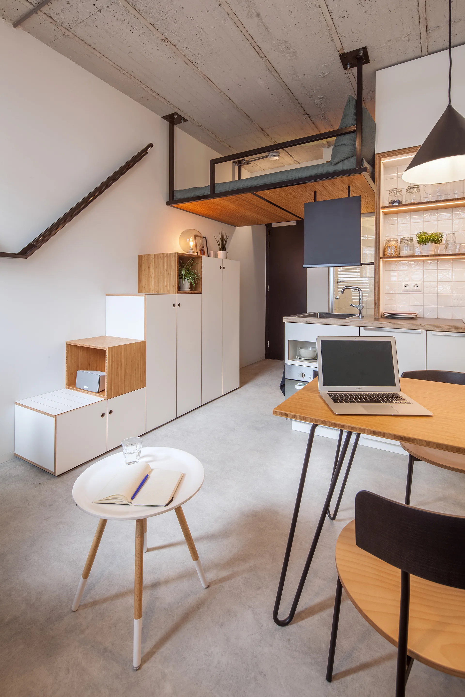 A compact studio apartment features a loft bed over the kitchen area, a concrete ceiling, and white wooden stairs that double as storage cabinets.