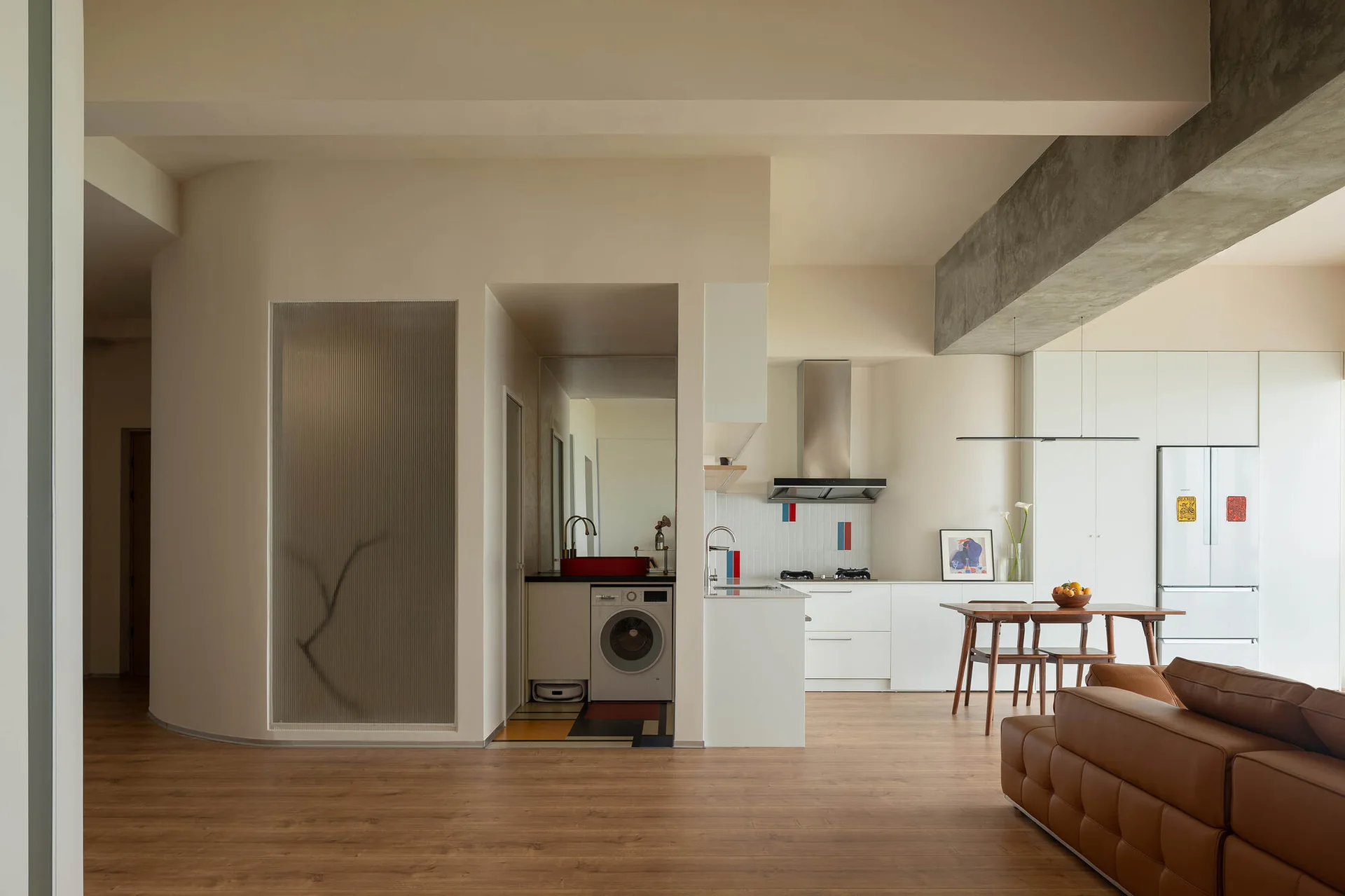 A modern minimalist kitchen and dining area featuring a curved partition wall with a fluted glass panel, a concrete ceiling beam, and a laundry nook.