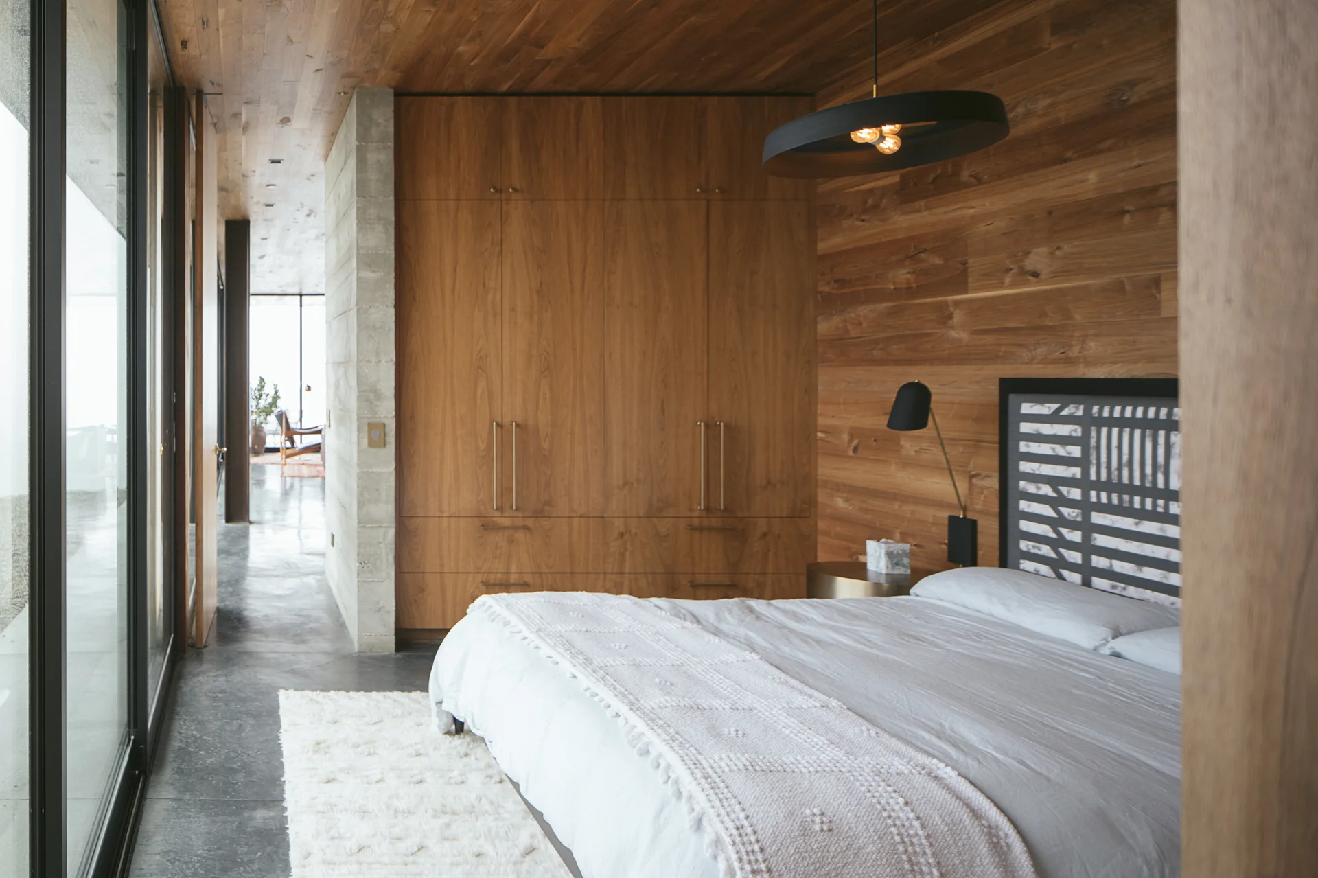 A modern bedroom featuring wood-planked walls and ceiling, a built-in wardrobe, a concrete partition wall, and glass sliding doors.