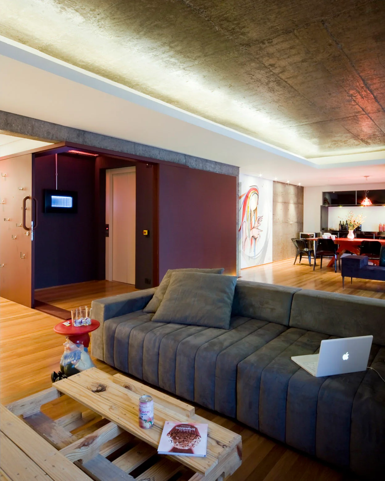 An industrial living room with a gray channel-tufted sofa, a pallet coffee table, light wood flooring, and an exposed raw concrete ceiling.