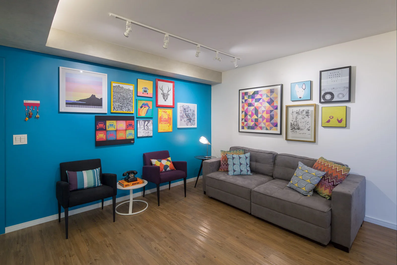 An eclectic living room features a gray sofa against a white wall and a bright blue accent wall decorated with a colorful gallery of framed art.