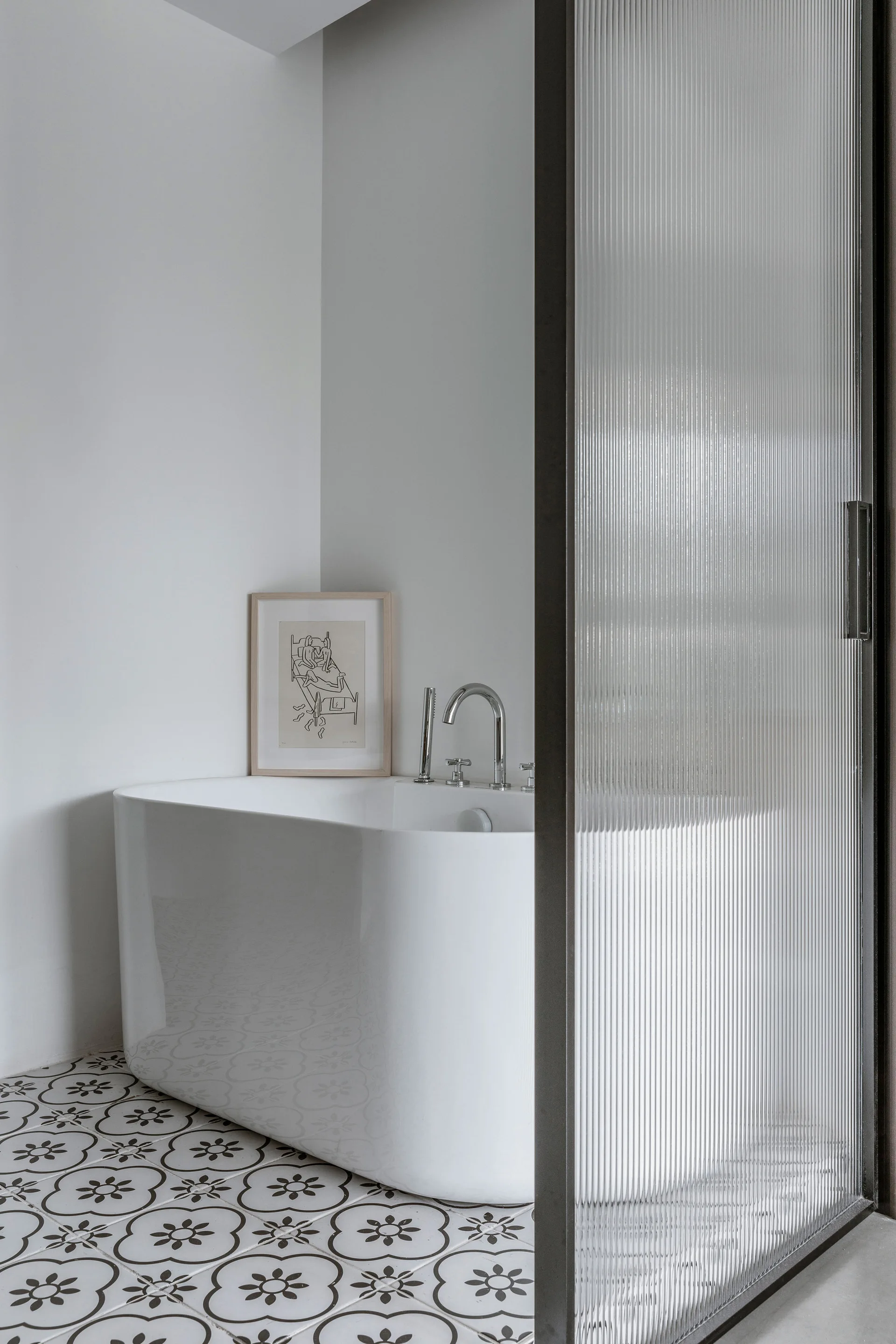 A white freestanding bathtub sits behind a reeded glass sliding door on patterned black and white tile flooring.