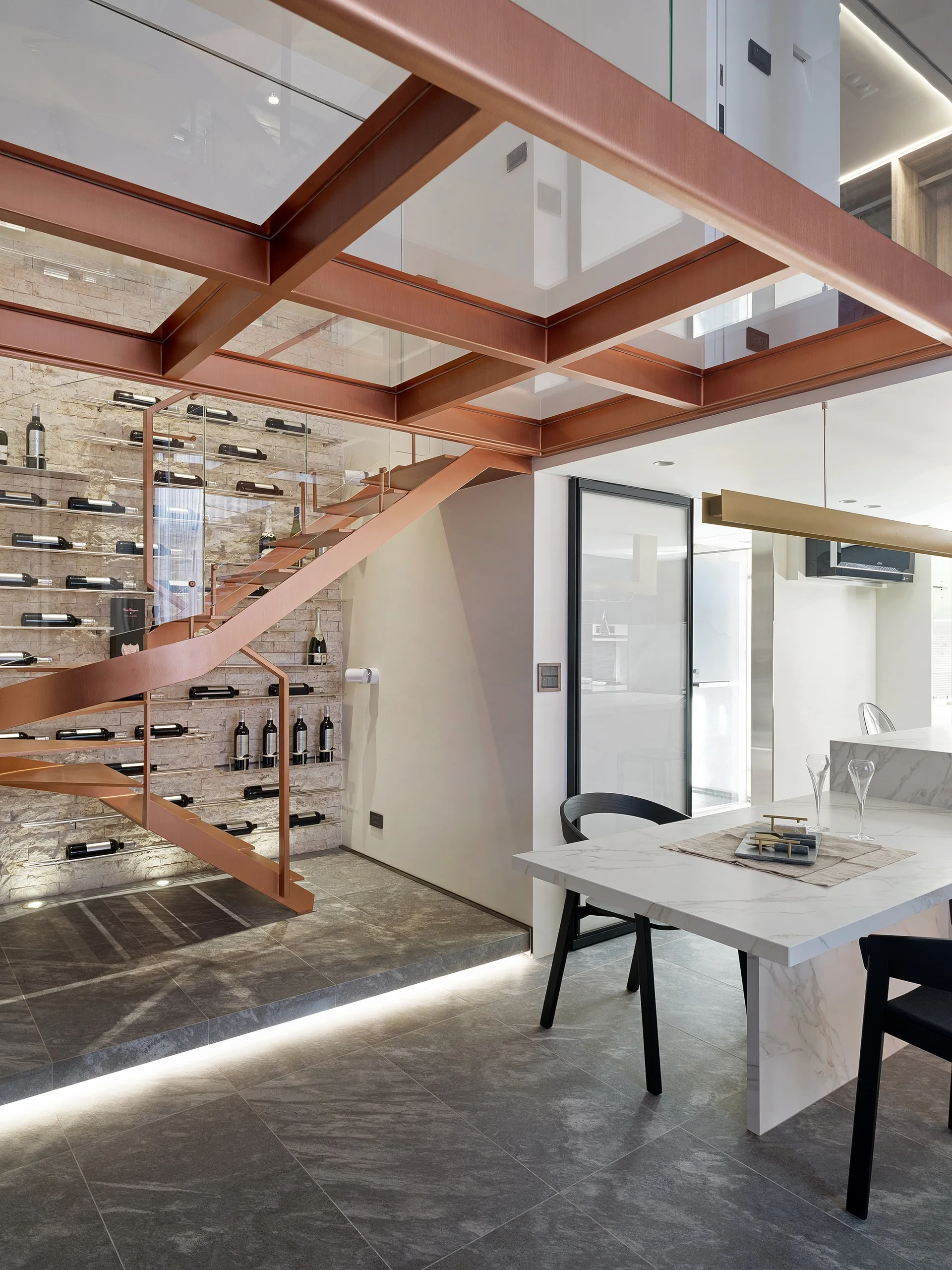 A copper spiral staircase rises above an under-stair wine display on a stone wall, next to a marble dining table with black chairs.