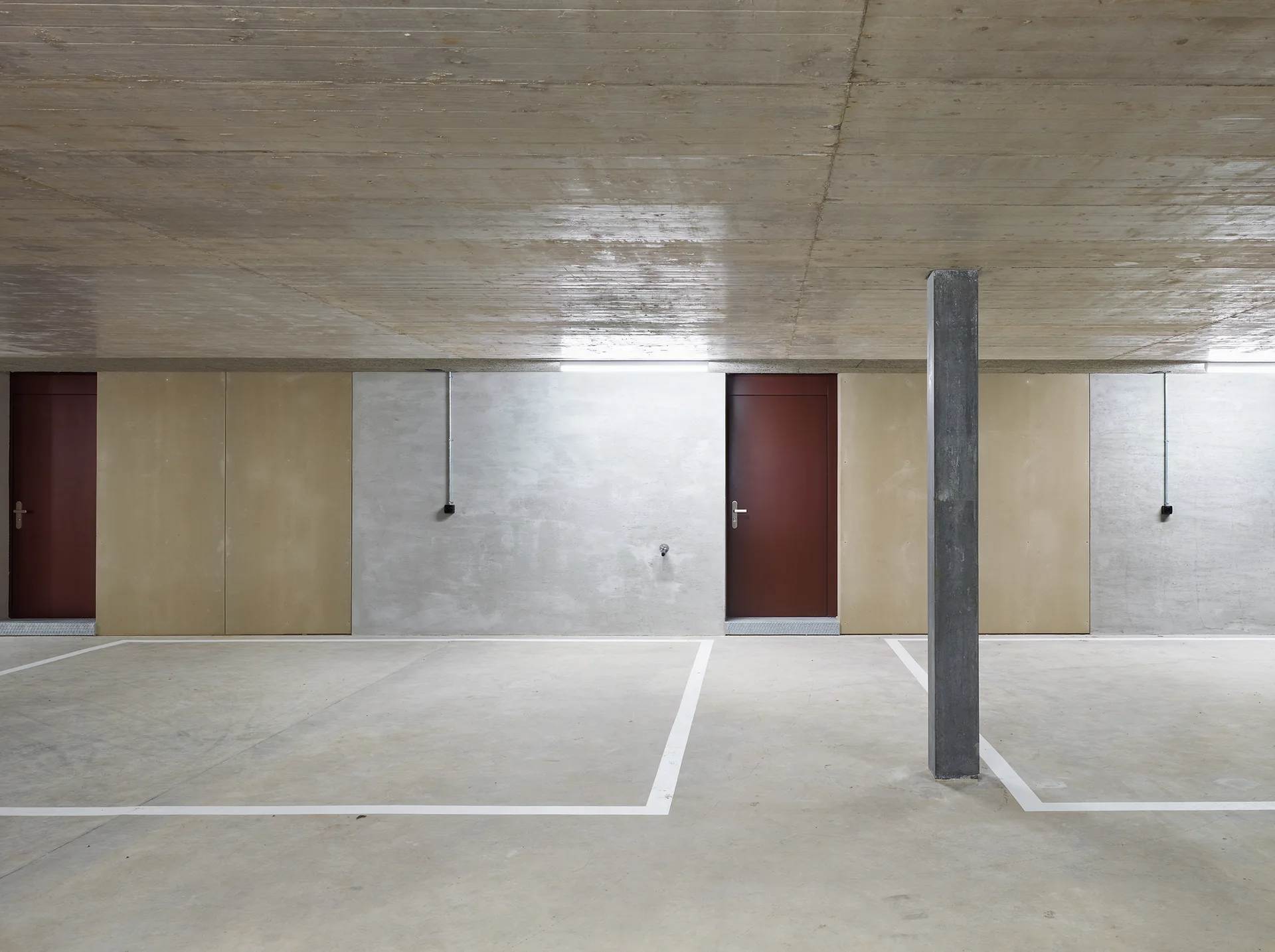 A concrete parking garage interior features a raw concrete ceiling, painted parking bays, a single vertical column, and two maroon doors.
