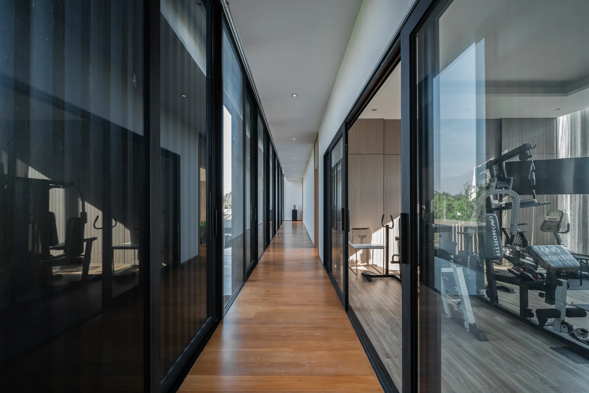 A long modern hallway features continuous wood plank flooring, white walls, and black-framed glass sliding doors that open to a home gym.