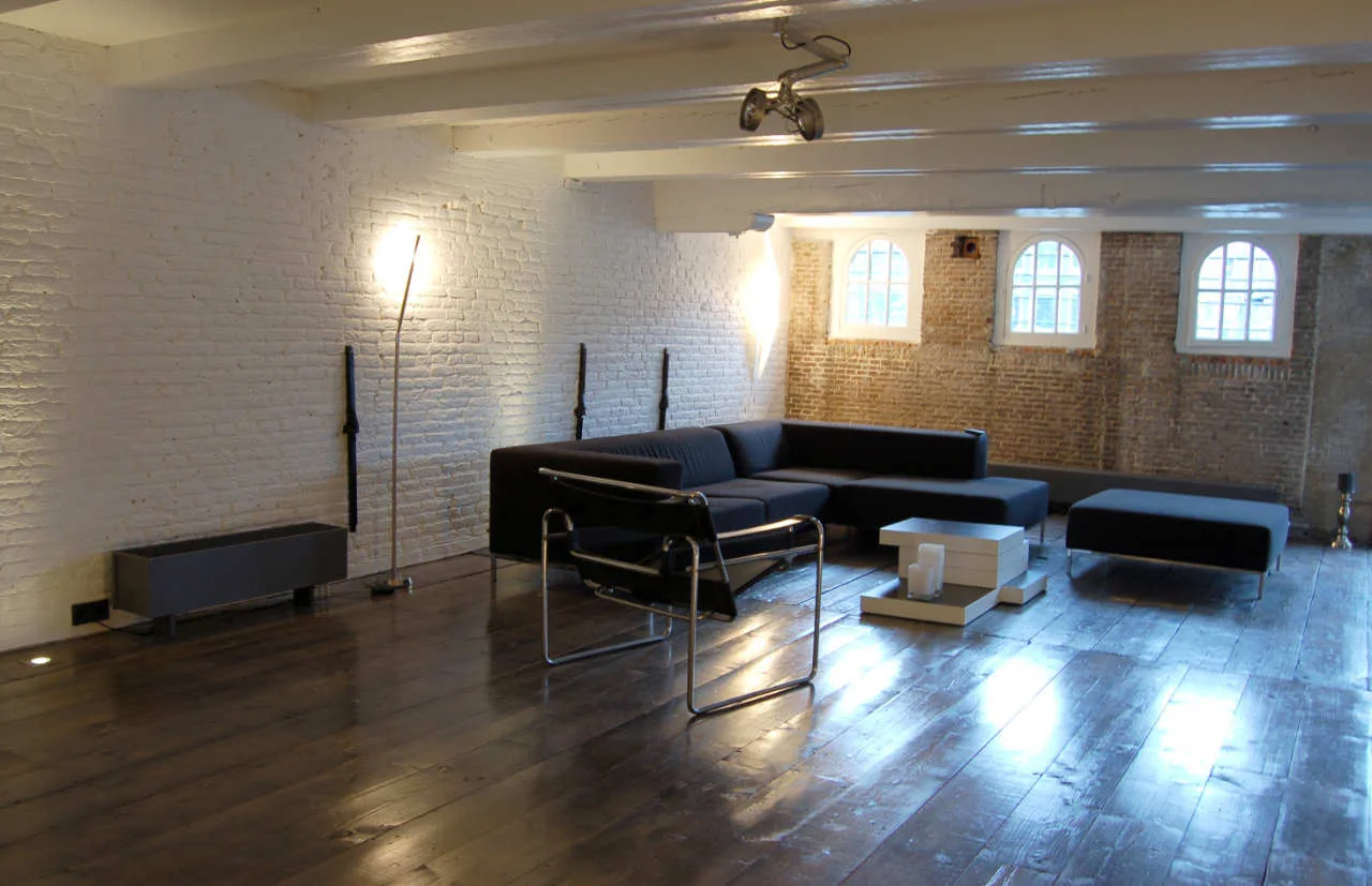 A minimalist loft living room features a low-profile black sectional sofa, a modernist armchair, glossy dark wood flooring, and exposed brick walls.