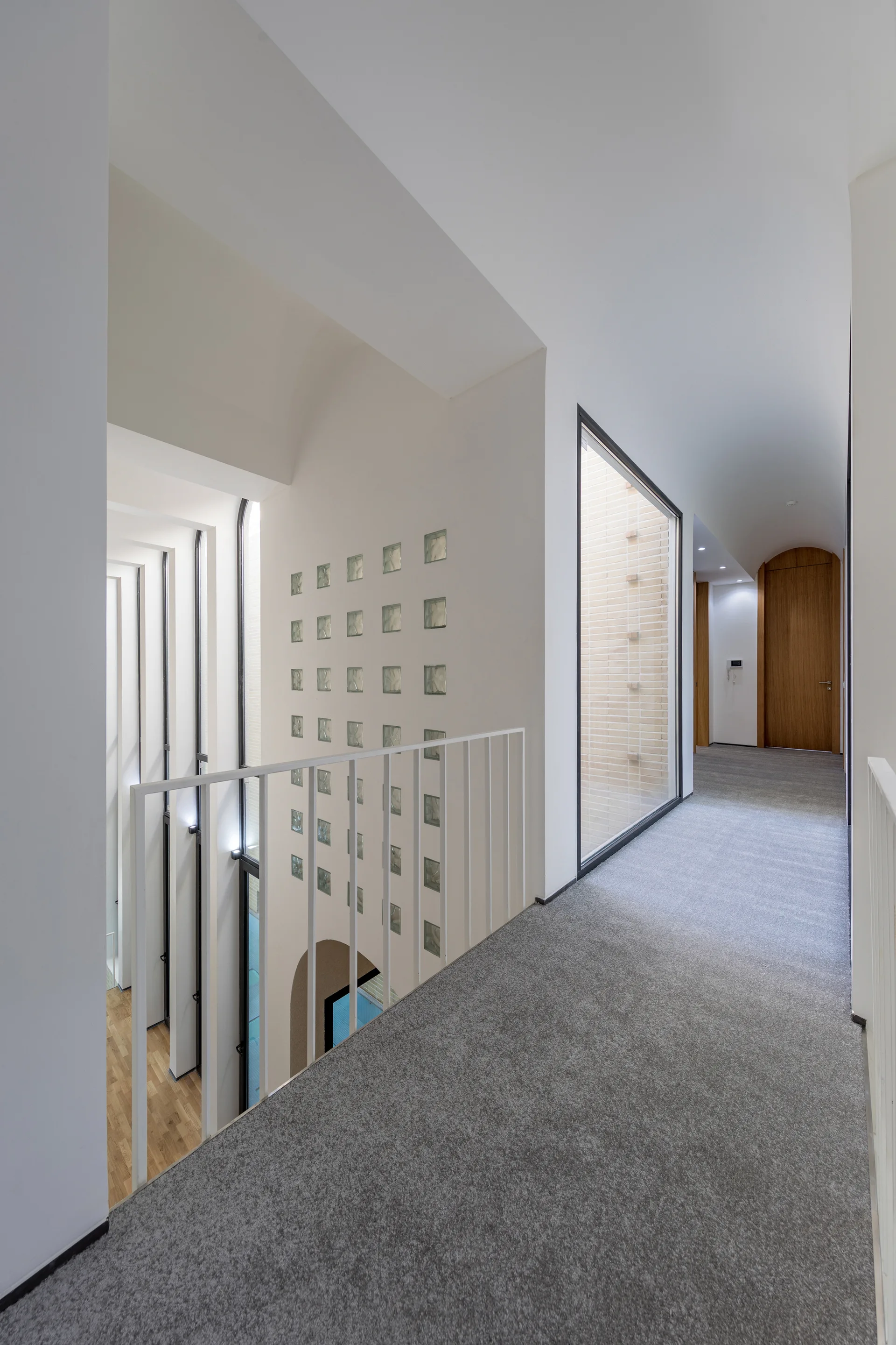 A modern hallway features gray carpet, a white metal railing, a large window, and a grid of glass blocks under a vaulted ceiling.