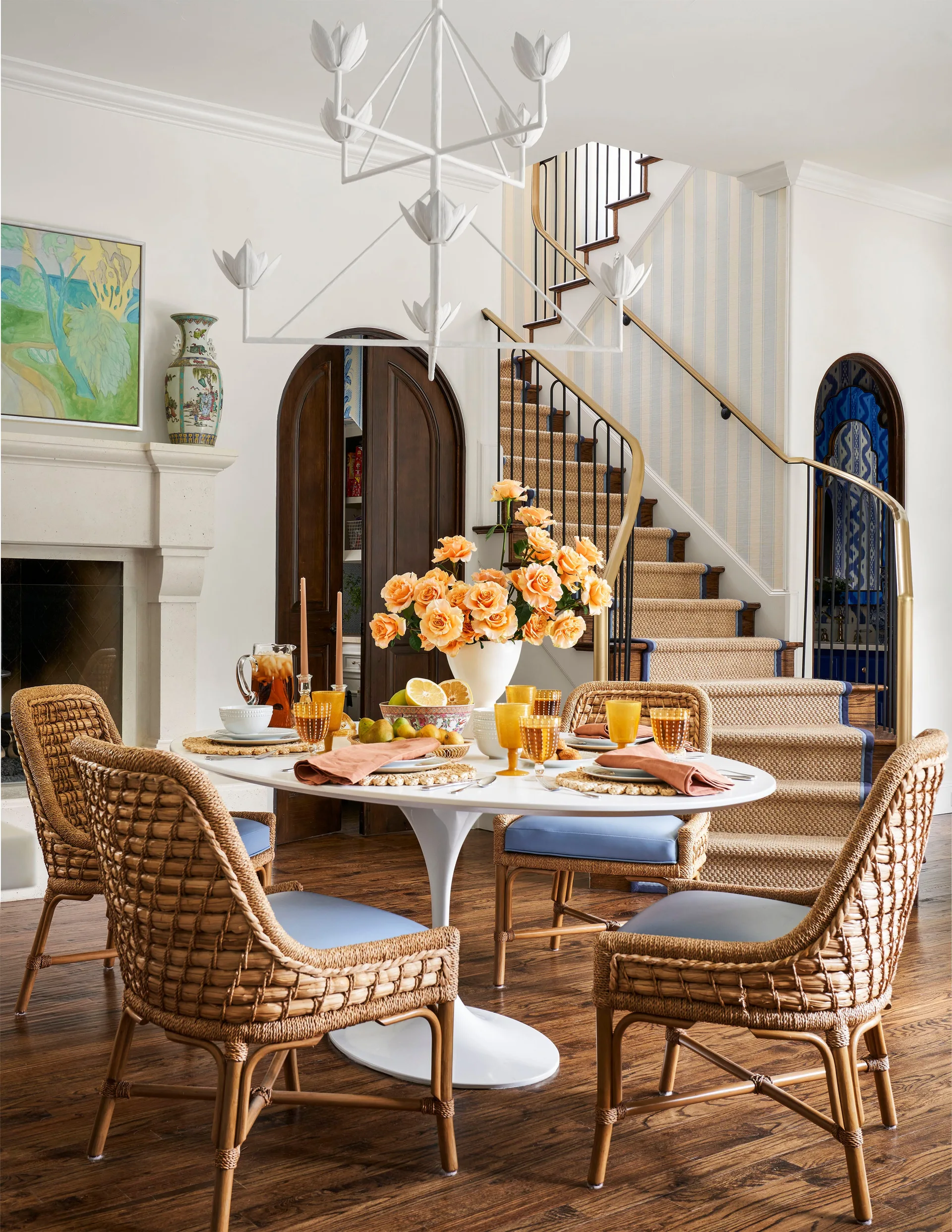 A white tulip table and woven wicker chairs with blue cushions sit in a bright dining room near a stone fireplace and staircase.