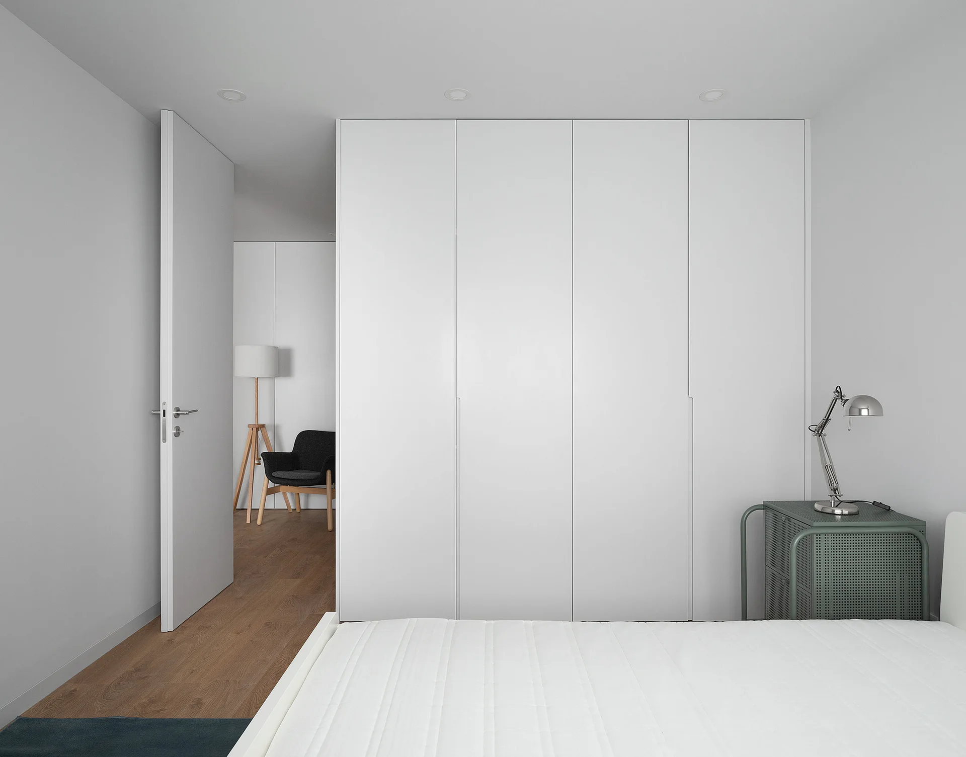 A minimalist white bedroom features a seamless floor-to-ceiling wardrobe, a green perforated metal side table, and an open doorway showing a second room.
