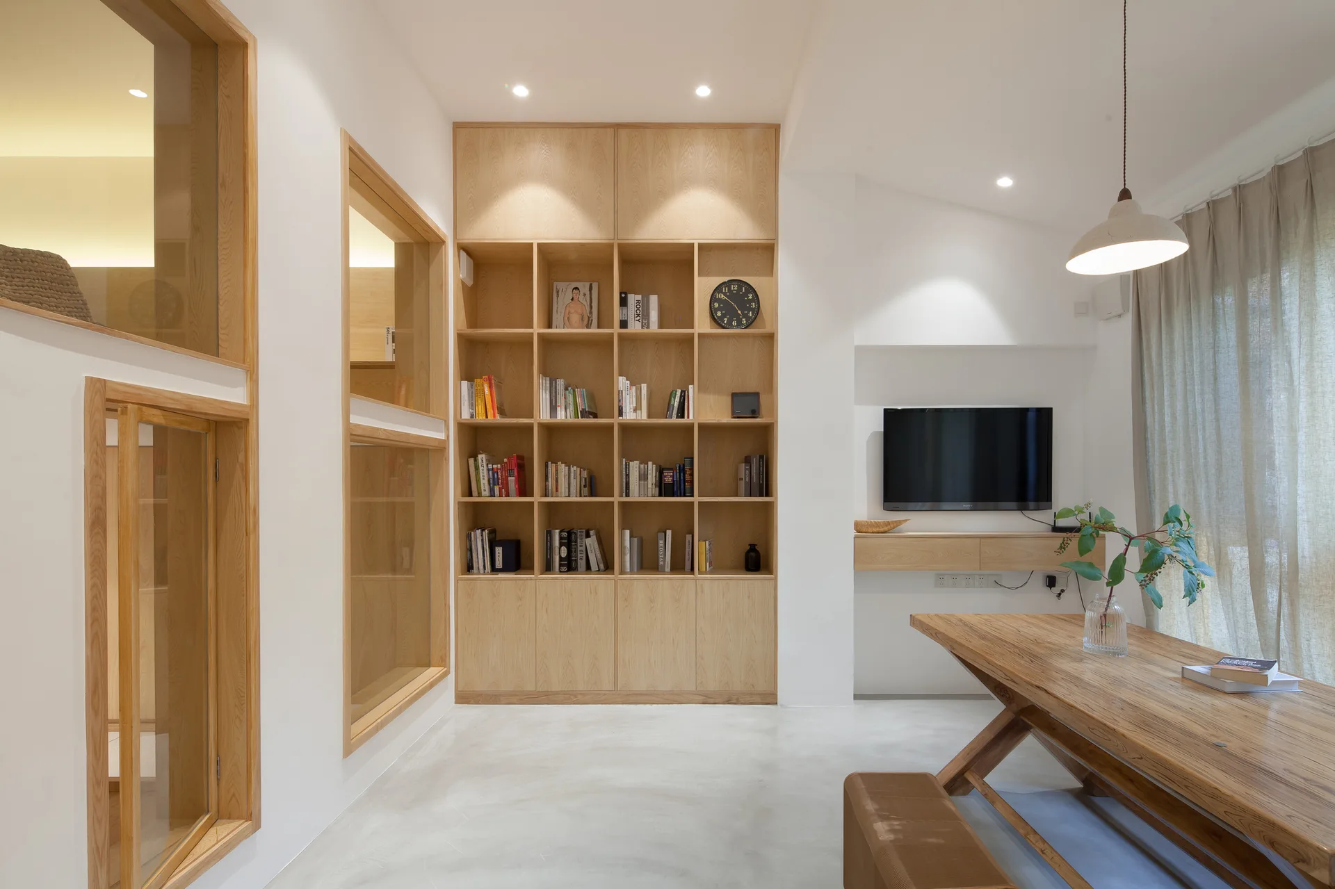 A minimalist room features custom light wood bookshelves, interior glass windows, and a long wooden dining table on concrete flooring.