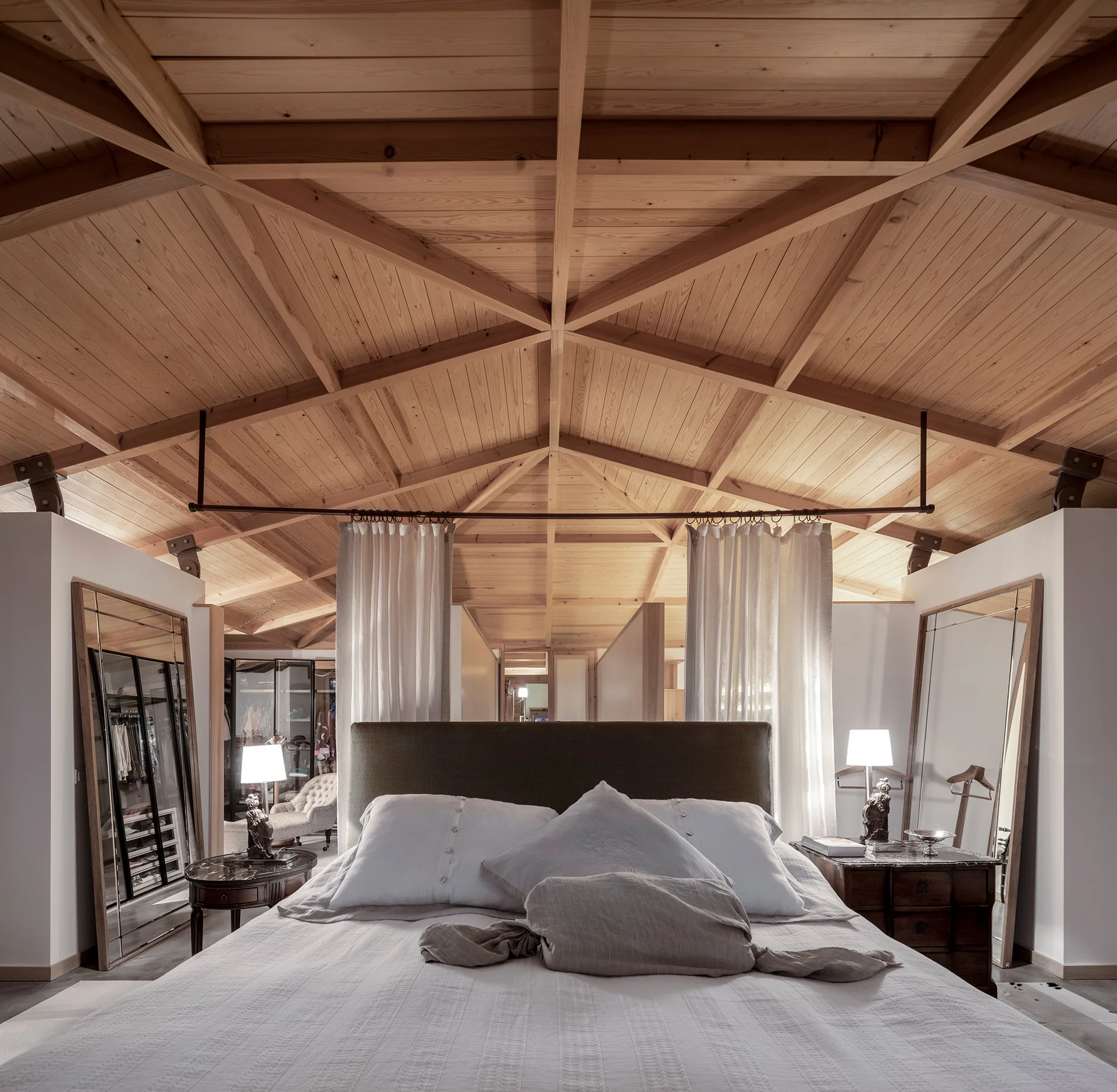 A bedroom features a high vaulted wood ceiling with exposed timber framing, a central bed with a dark headboard, and flanking standing mirrors.