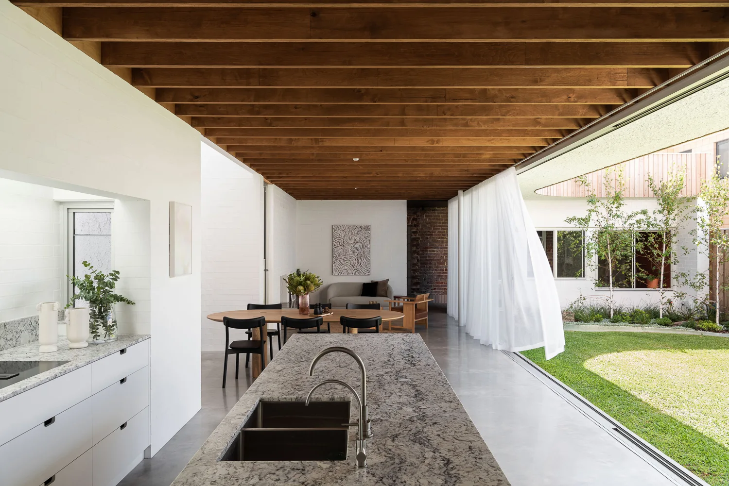 An open kitchen and dining area features an exposed timber ceiling, a stone-topped island, and a sheer curtain opening to an outdoor lawn.