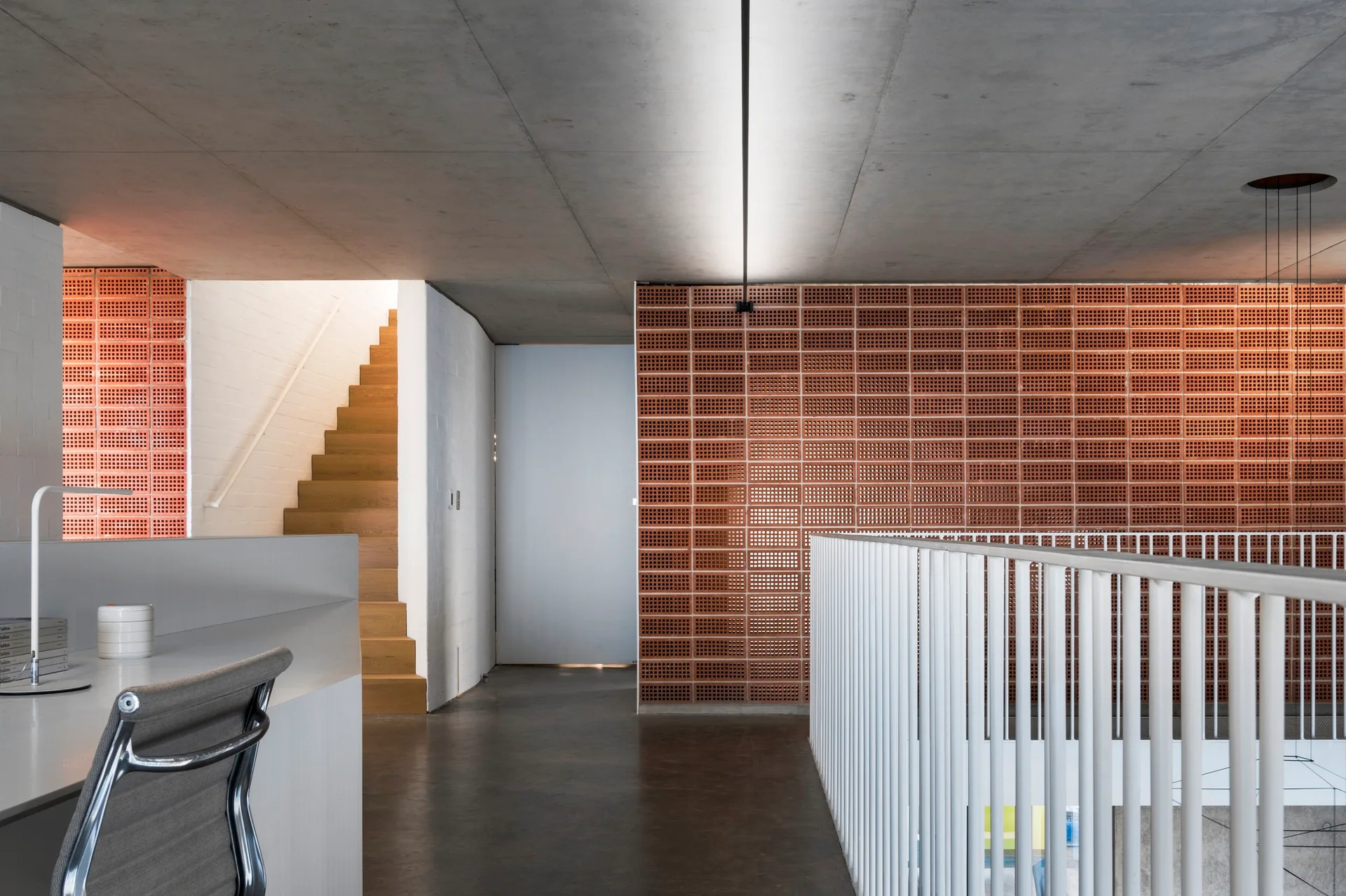 A modern interior landing features a terracotta block screen wall, a smooth concrete ceiling, a wooden staircase, and a white metal railing.