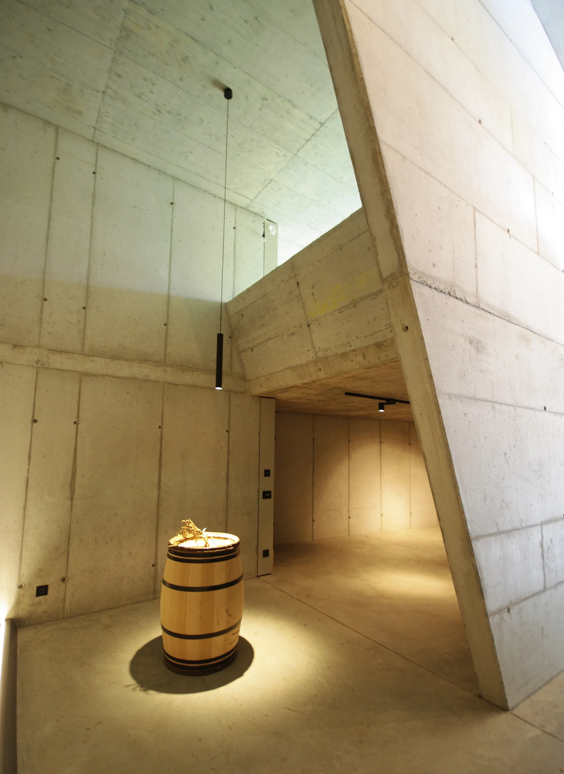 A minimalist architectural space with exposed board-formed concrete walls, a smooth concrete floor, and a single illuminated wooden barrel in the center.
