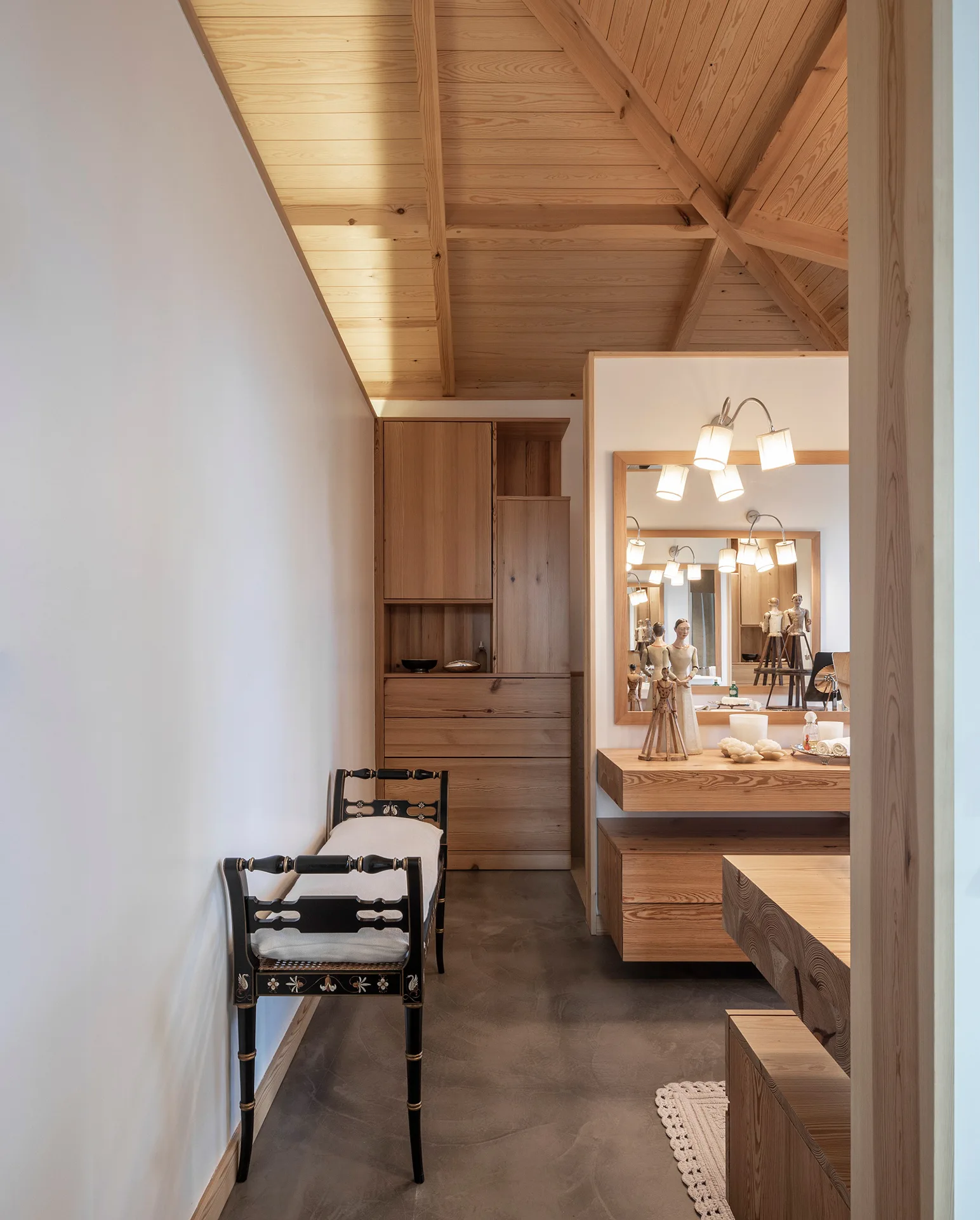 A light-filled dressing area features a floating wooden vanity, a vaulted wood plank ceiling, a smooth concrete floor, and a black painted bench.