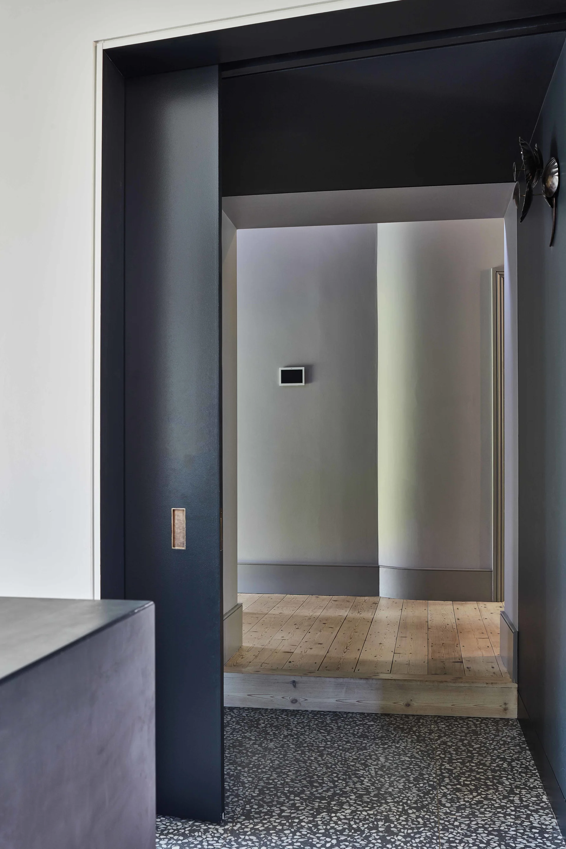 A dark charcoal sliding door frames a transition from speckled grey terrazzo flooring to a light wood floor hallway.