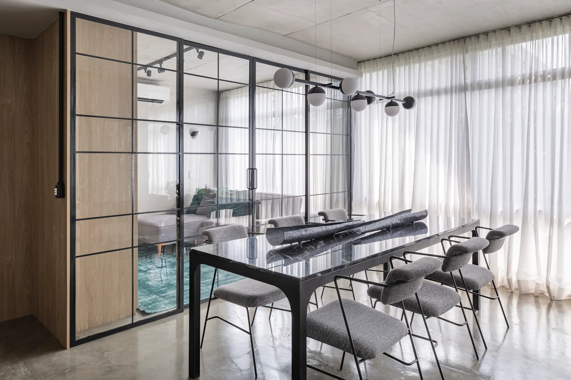 A modern dining room features a black stone table and grey chairs next to a black-framed glass partition wall.