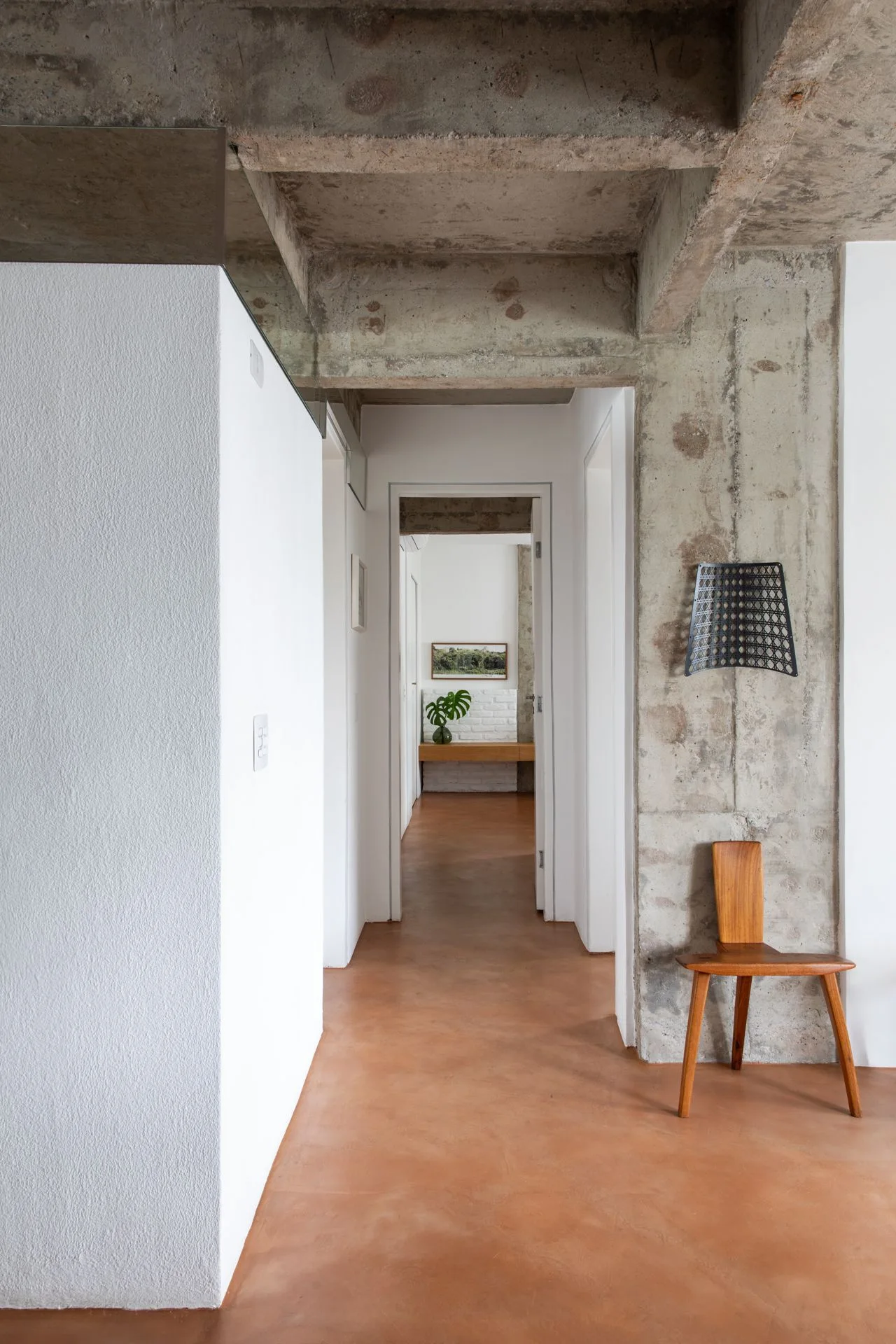 A minimalist hallway features terracotta-colored flooring, white plastered walls, and an exposed concrete ceiling with a wooden chair resting against a column.