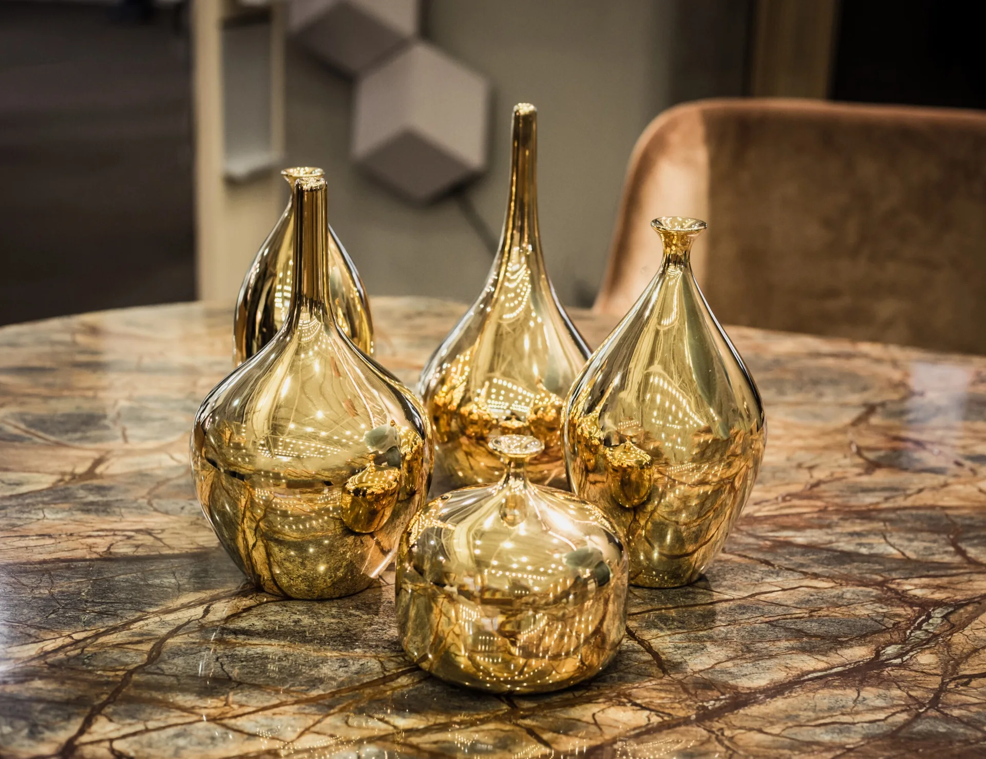 A collection of reflective gold metallic vases and a small jar rest on a brown veined marble tabletop.