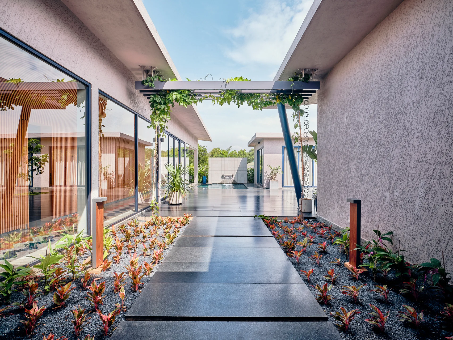 A central dark stone walkway leads through a modern gravel courtyard with low red-green plants, glass walls, and an overhead vine-wrapped pergola.
