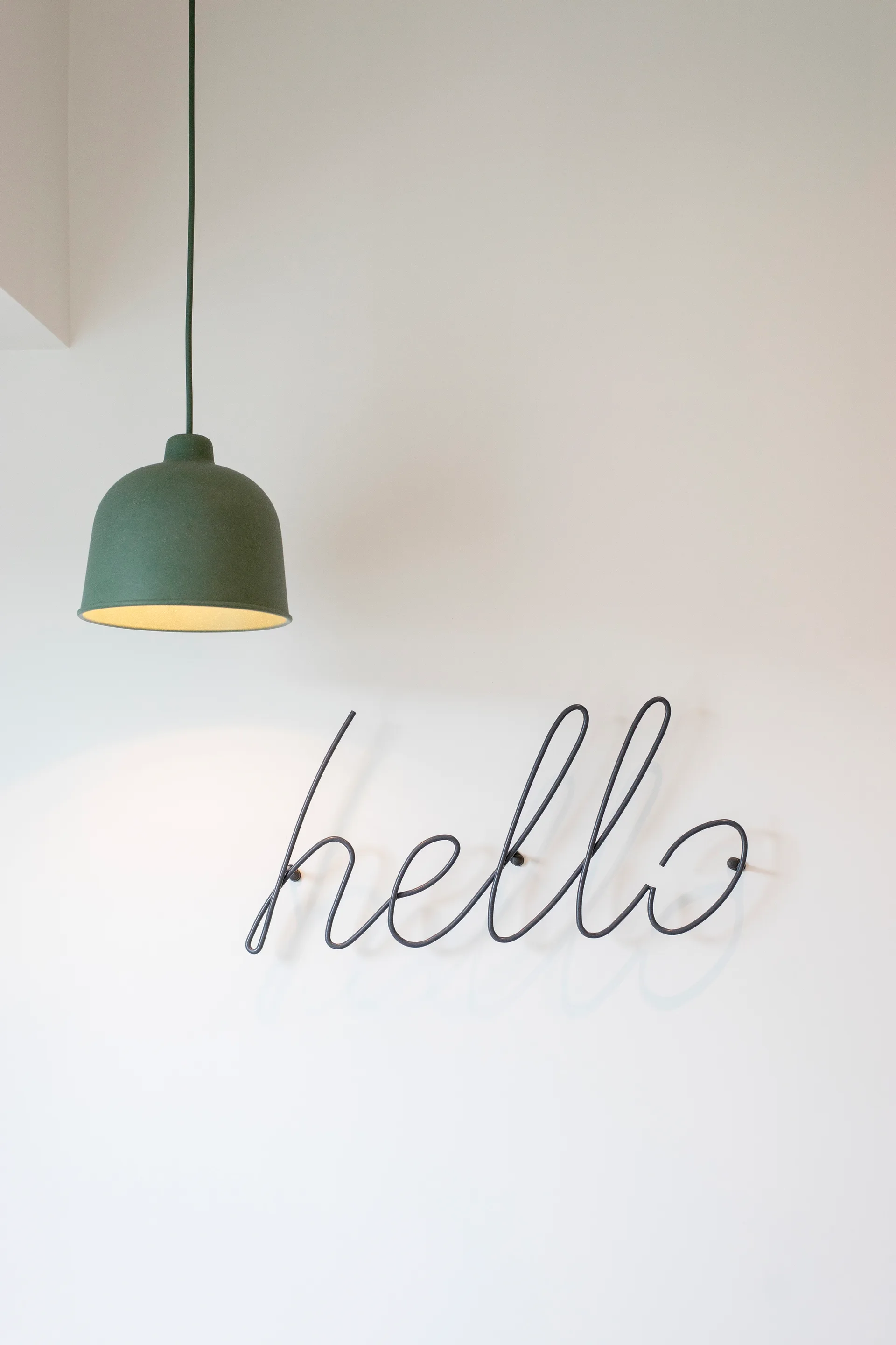 A matte green dome pendant light hangs above a black cursive metal sign spelling hello on a plain white wall.
