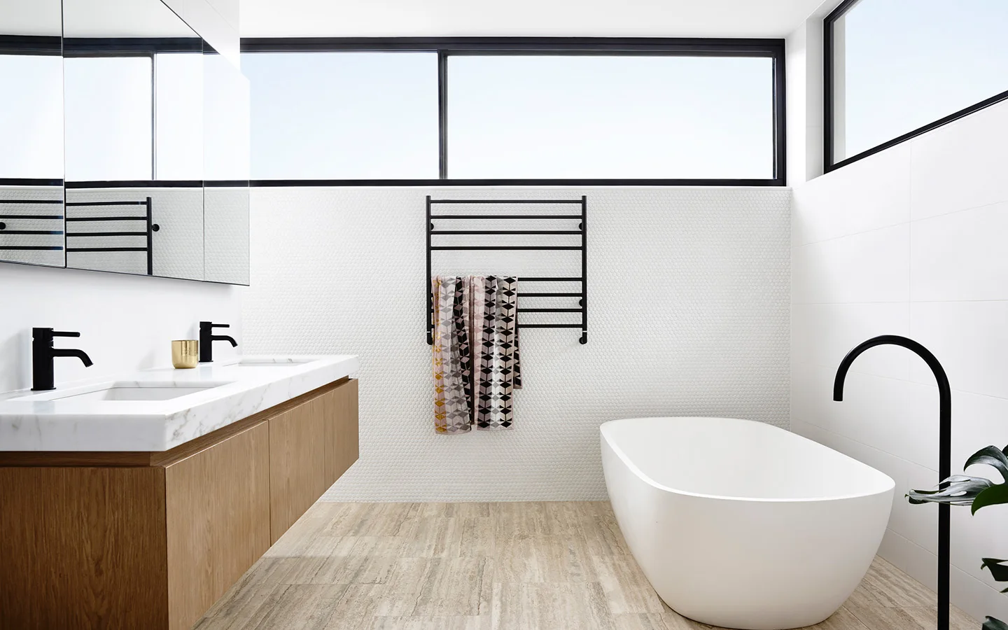 A modern bathroom featuring a floating wood vanity, white freestanding bathtub, matte black fixtures, and white penny tile walls.