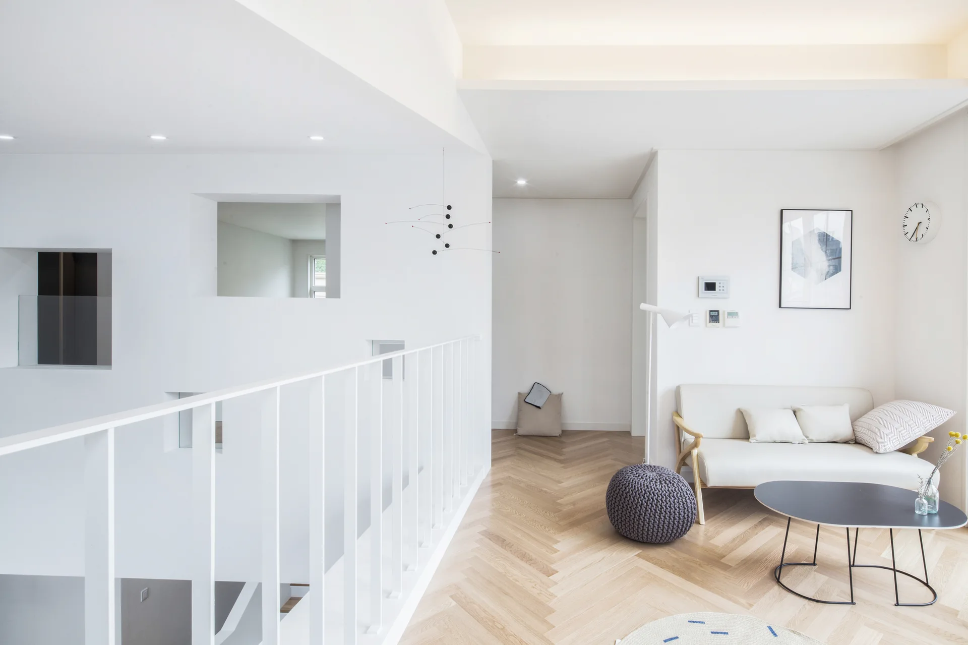 A minimalist loft living room features a cream sofa, a black coffee table, light wood herringbone floors, and a white metal staircase railing.